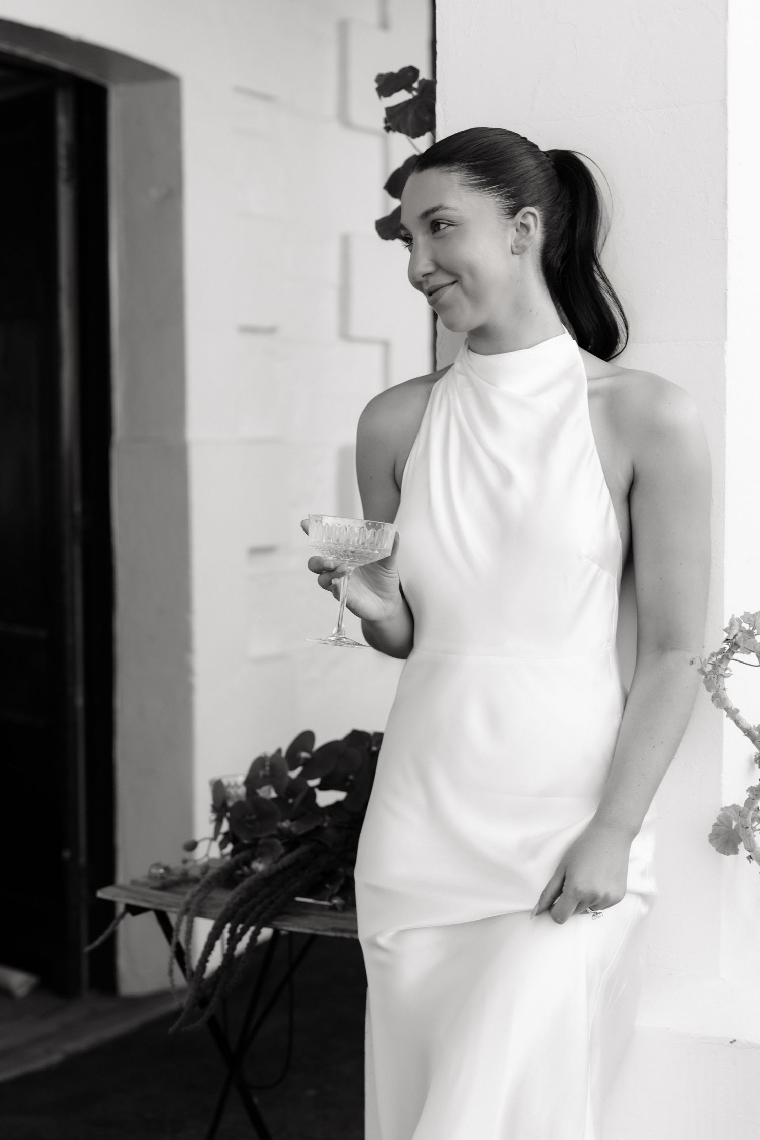Elegant bride in white halter gown holding champagne glass, black and white wedding photography.