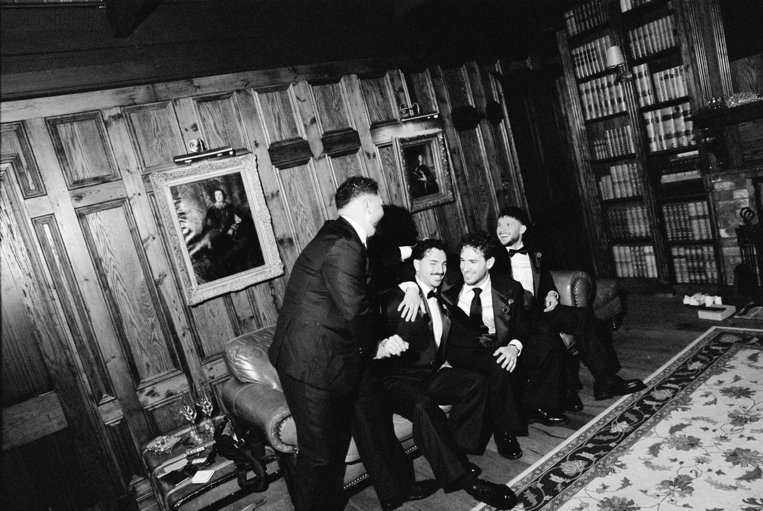 Group of men in tuxedos socializing in wood-paneled library at formal wedding reception.