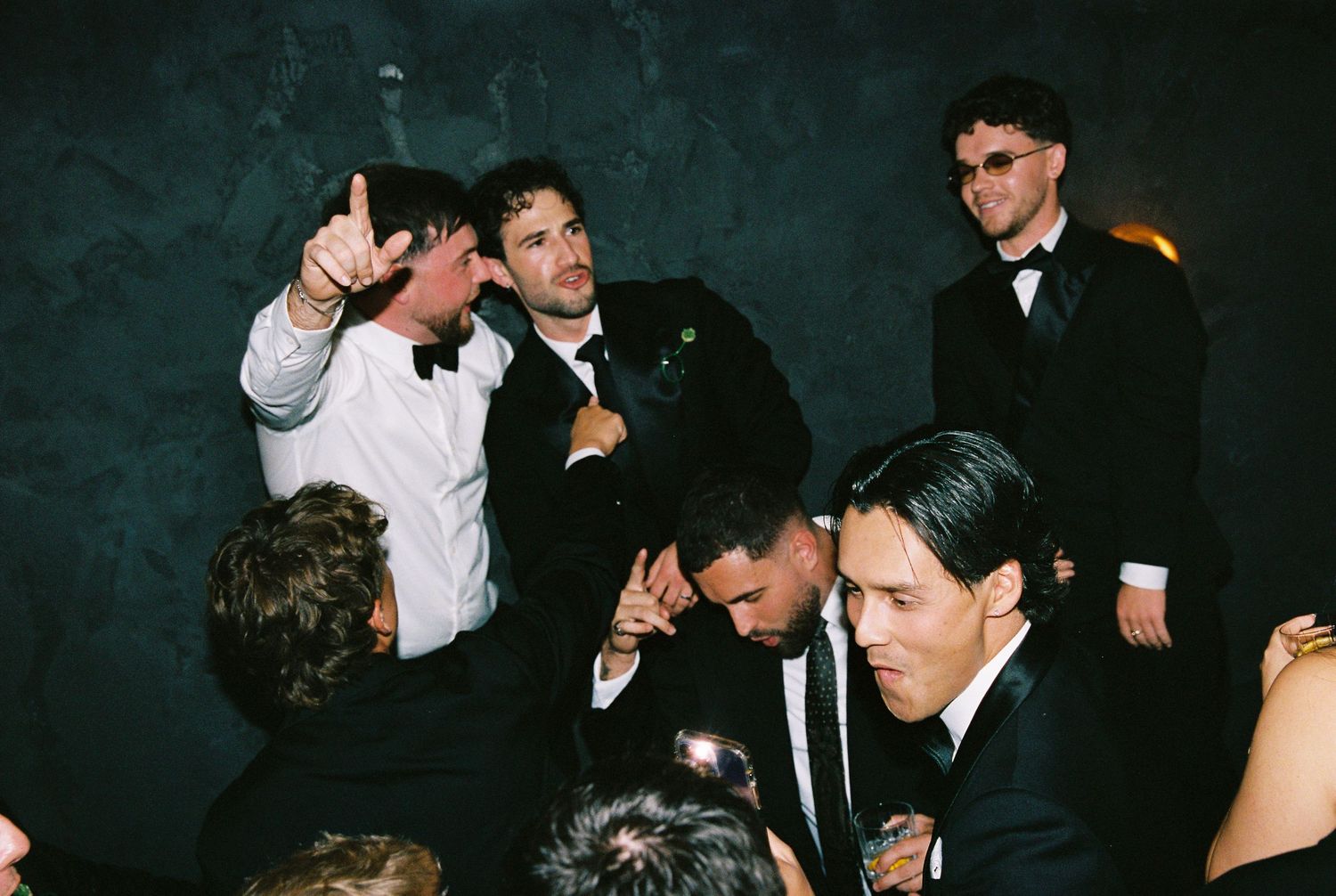 Group of men in formal suits celebrating and dancing energetically at a wedding reception.