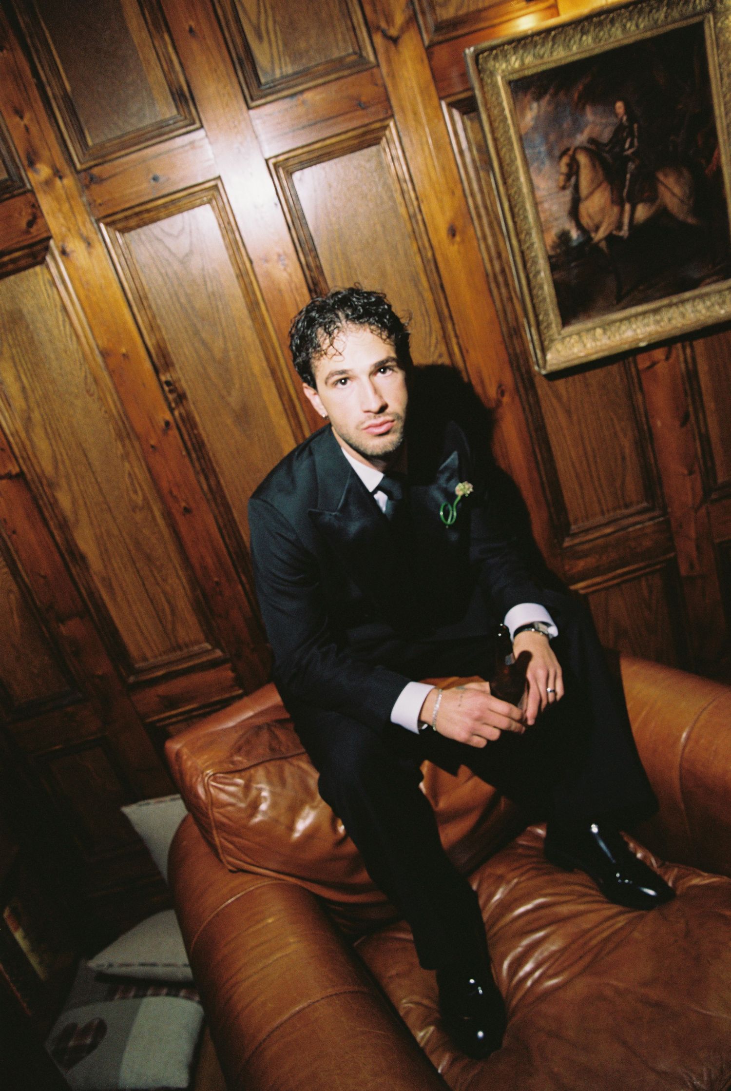 Groom in navy suit sits on leather sofa in wood-paneled room with equestrian portrait.