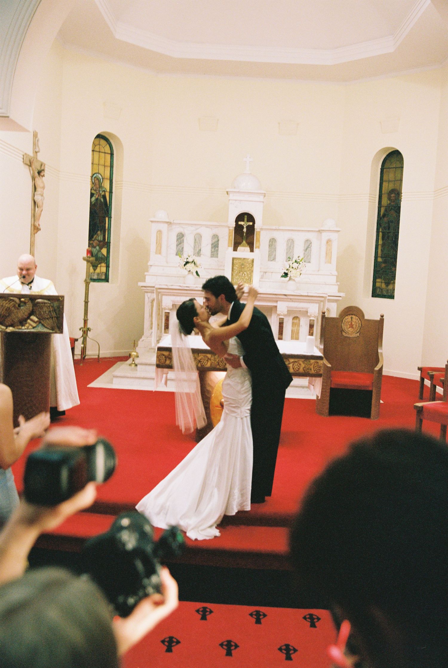 Bride and groom share first kiss at church altar with red carpet and stained glass windows.