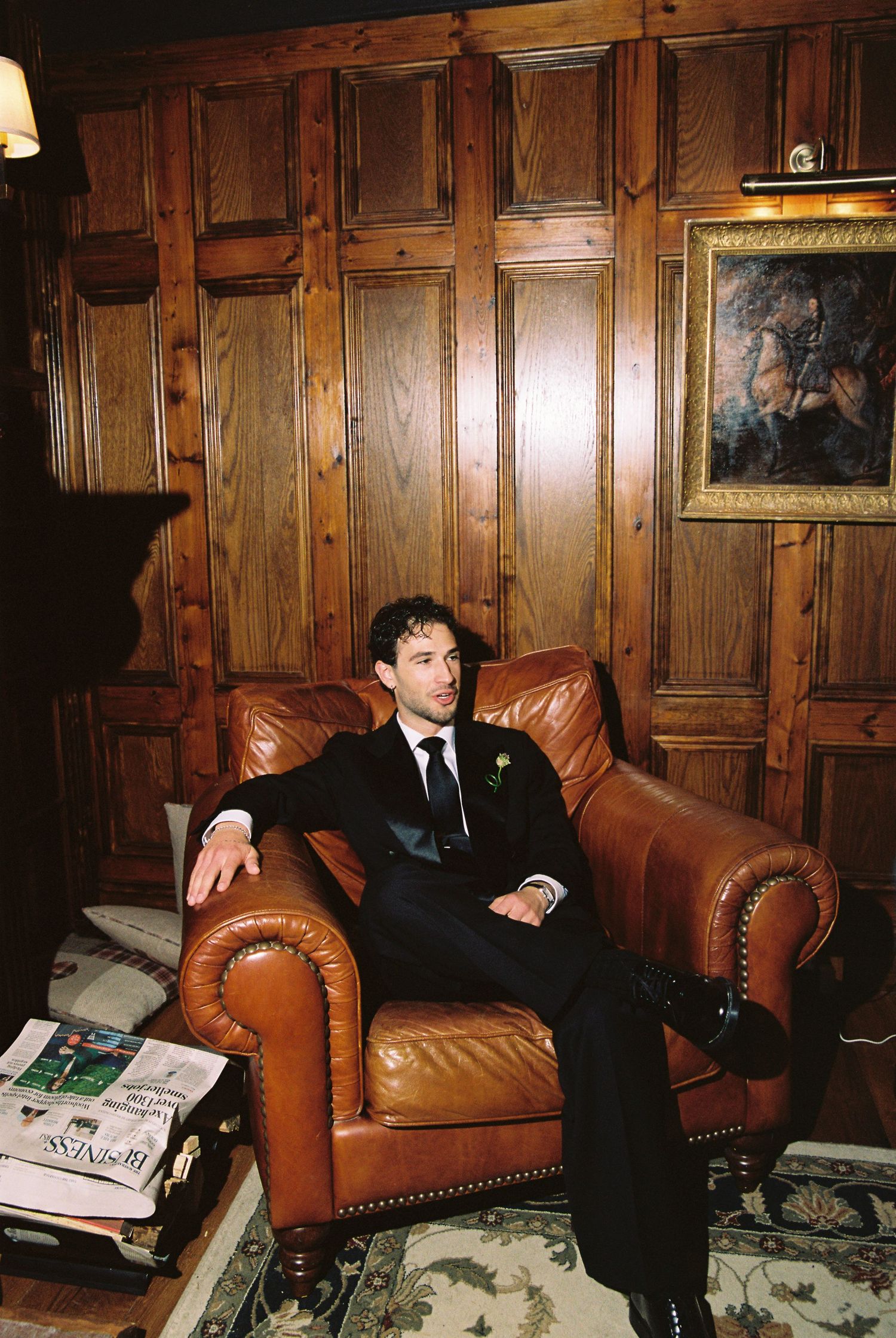 Man in black suit sitting relaxed in leather armchair in elegant wood-paneled room with painting.