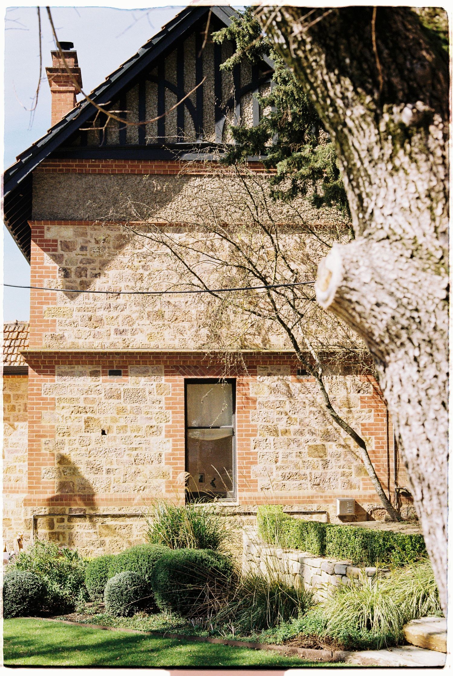 Rustic sandstone and brick heritage building surrounded by manicured gardens and mature trees in sunlight.