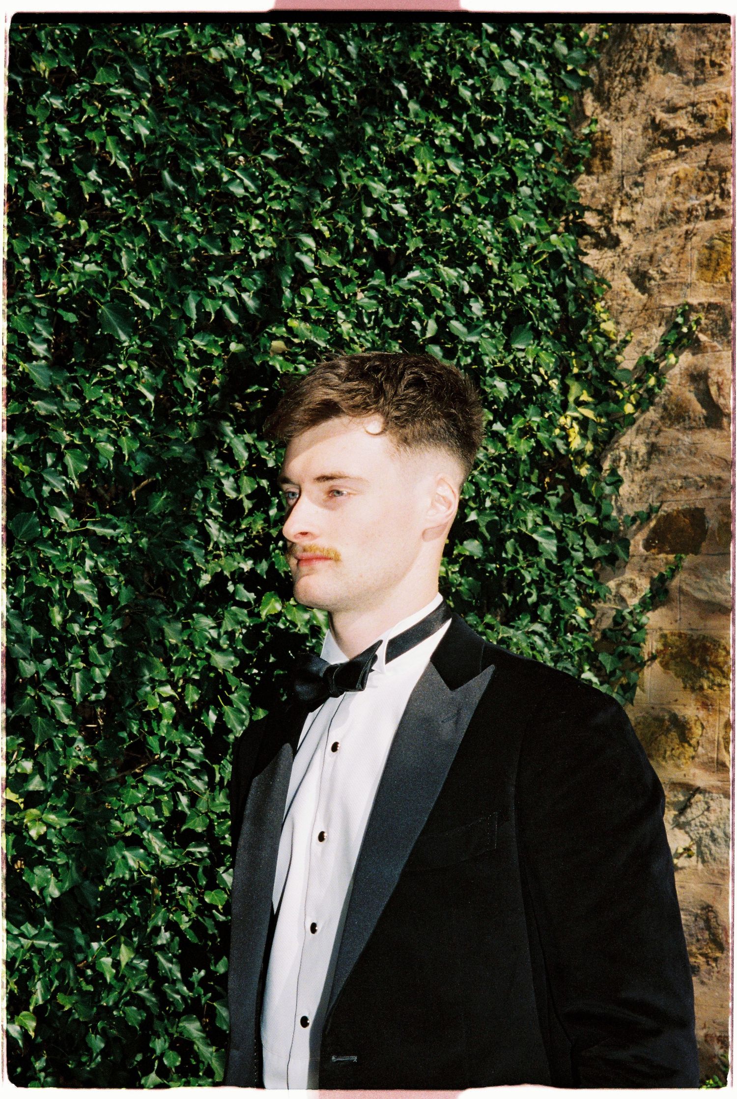 Groom in black tuxedo with bow tie standing against a lush green ivy-covered stone wall.