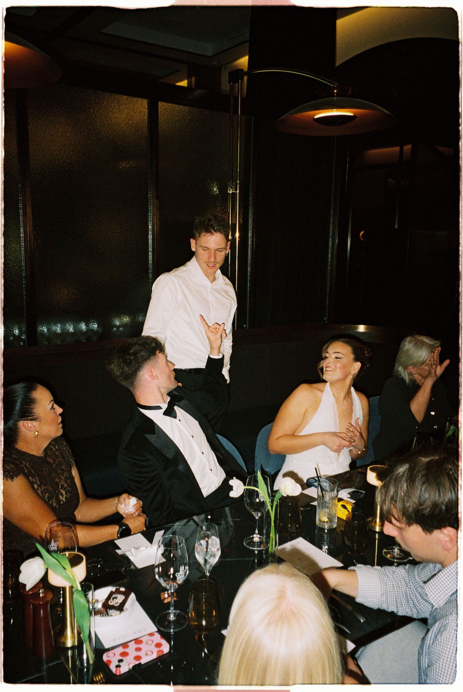 Wedding guests laughing and celebrating at a dark elegant reception dinner with a standing guest speaking.