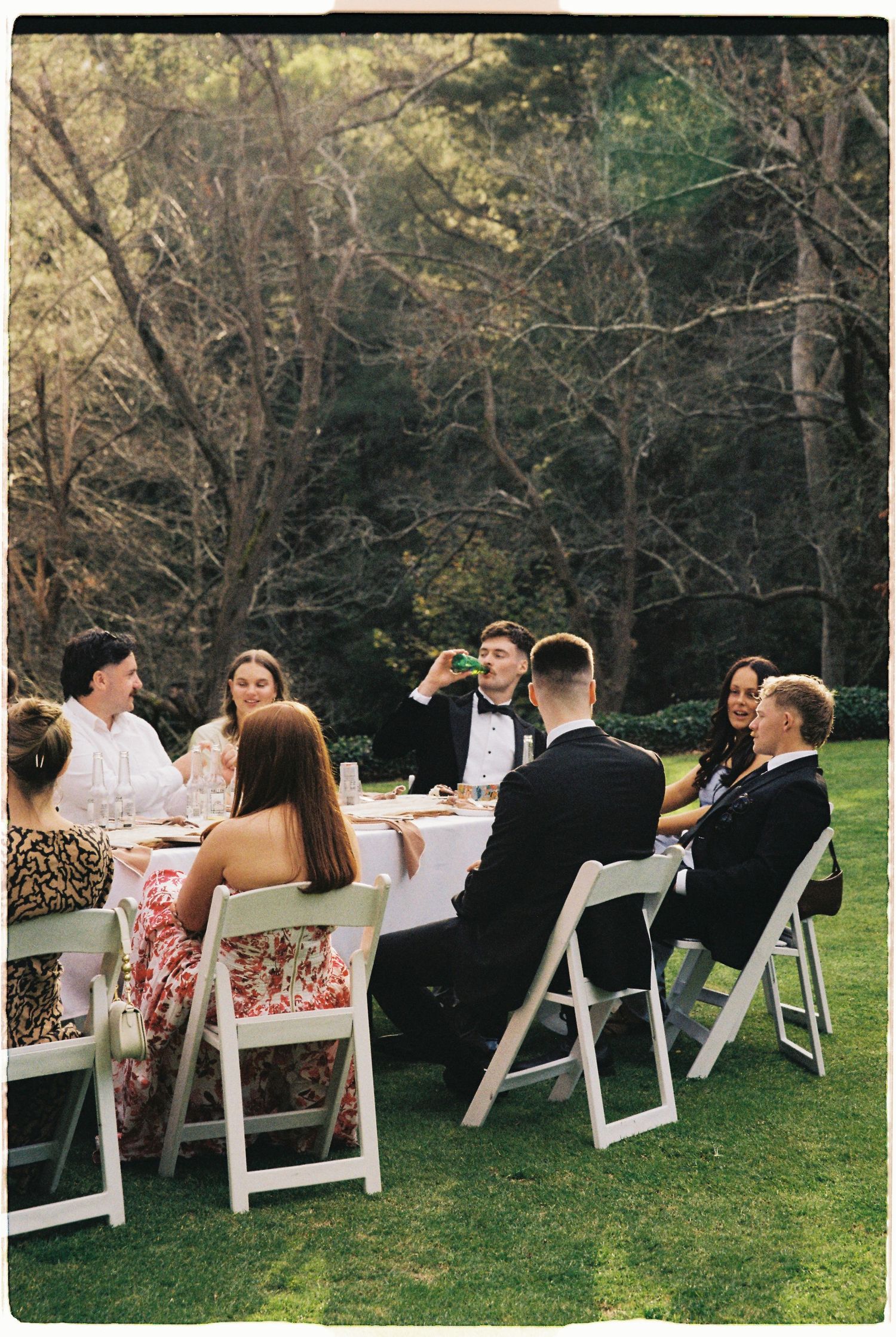 Wedding guests in formal attire enjoy outdoor reception dinner at white tables on green lawn.