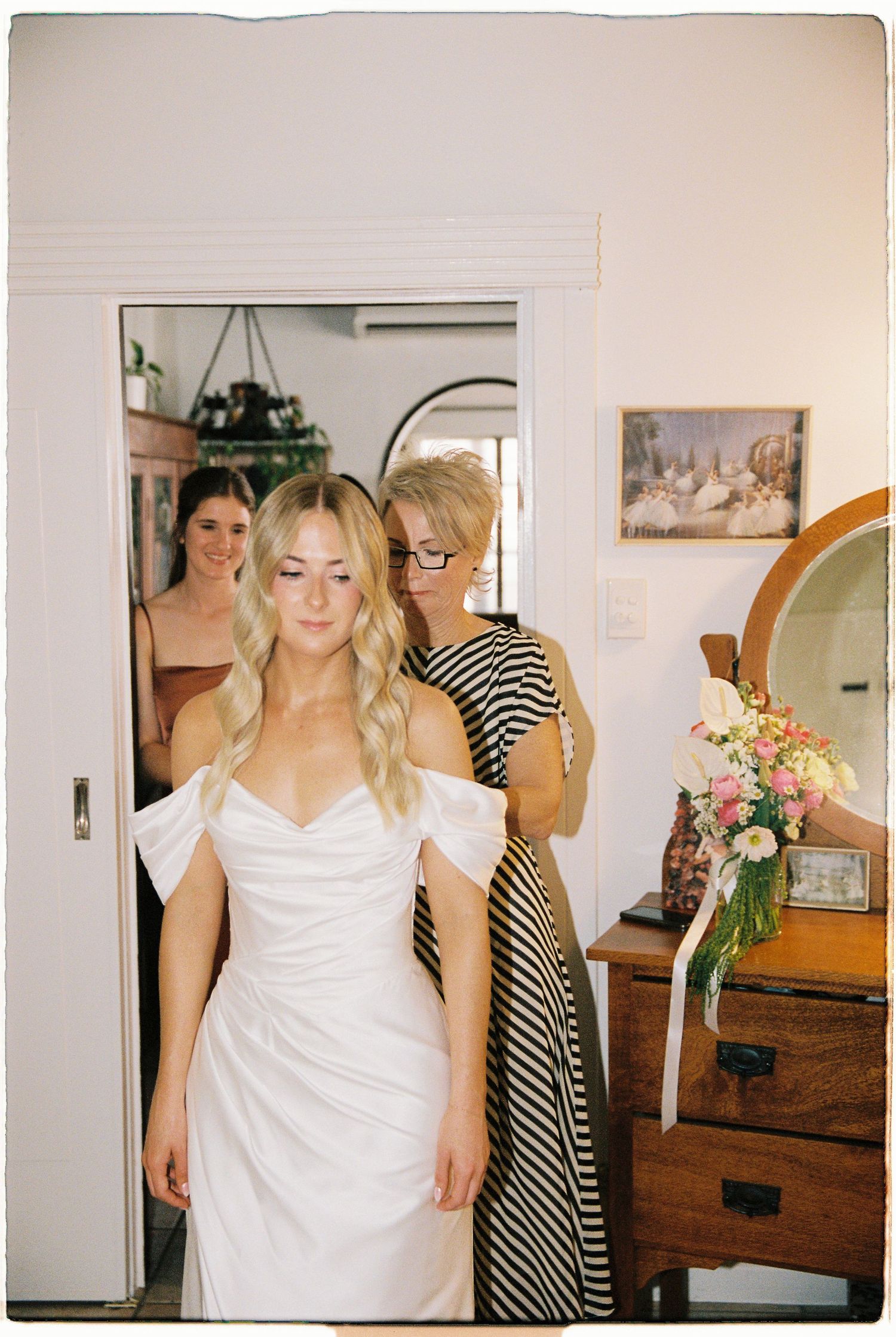 Bride in white off-shoulder gown getting ready with help from mother and bridesmaid in doorway.
