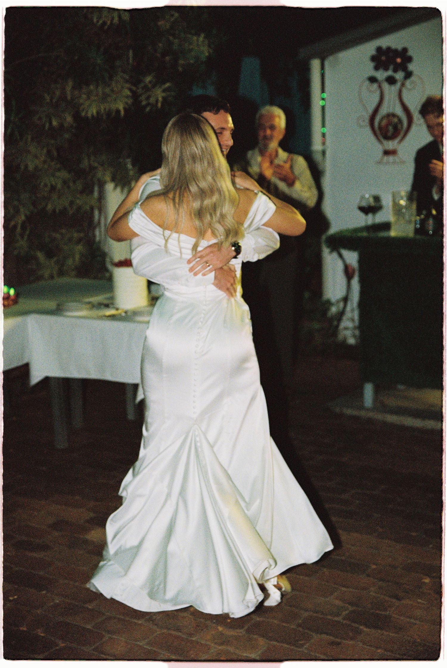 Bride in elegant white satin off-shoulder gown shares first dance with groom at outdoor wedding.