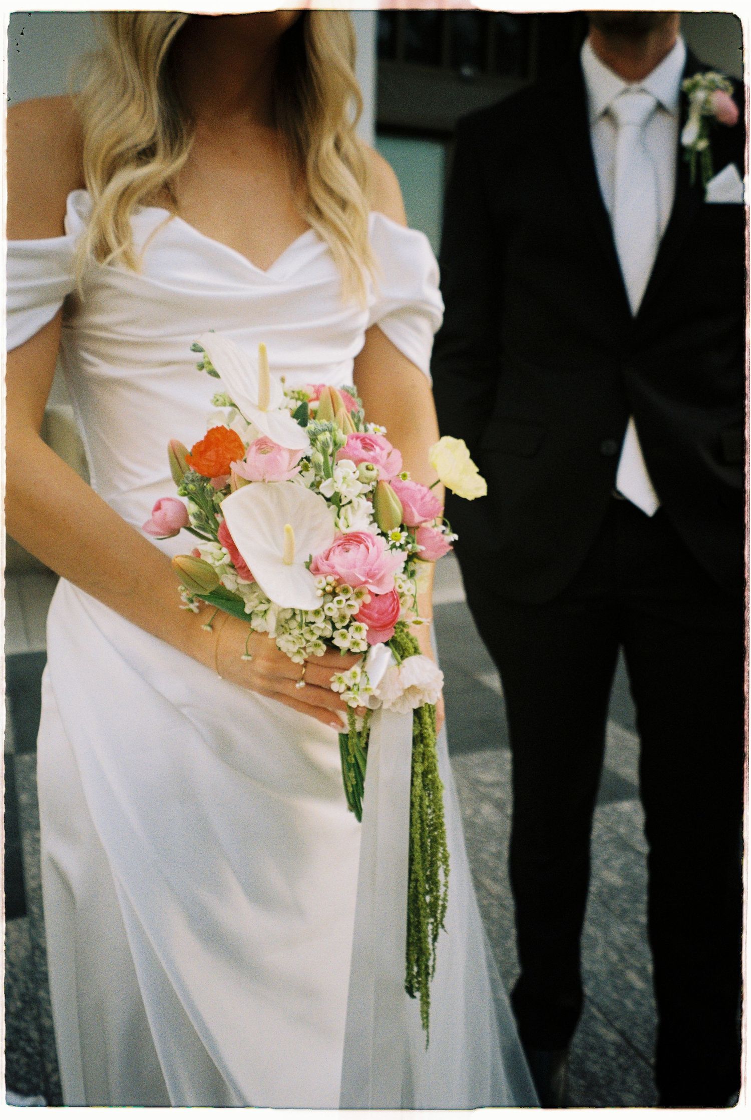 Blonde bride in off-shoulder white gown holds colorful pink and white bouquet beside groom in black tuxedo.
