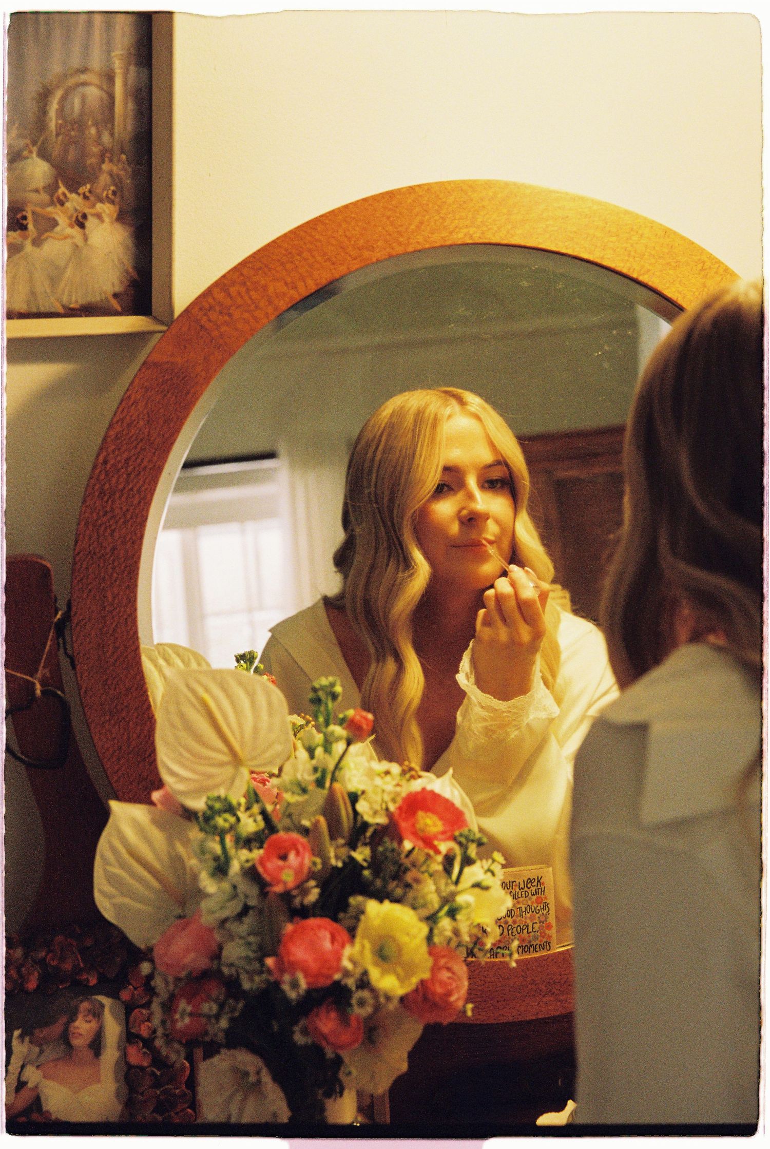 Bride applying makeup at round mirror surrounded by colorful fresh floral arrangement.