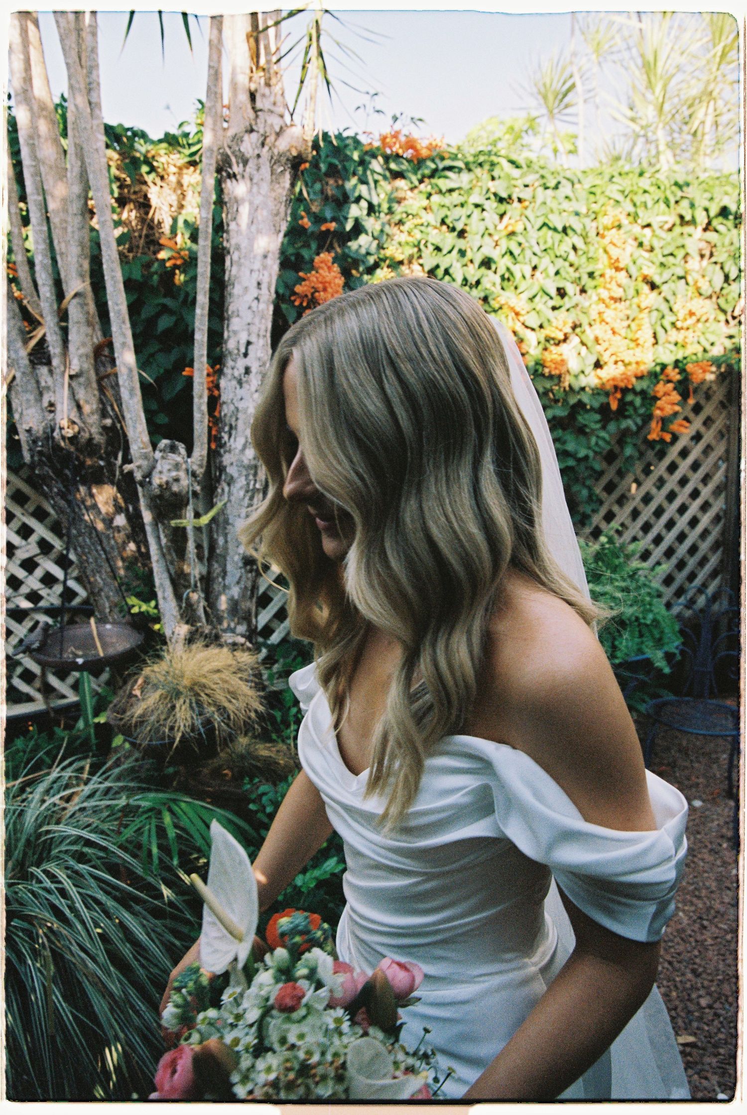 Blonde bride in off-shoulder white gown holding bouquet in sunlit garden setting.