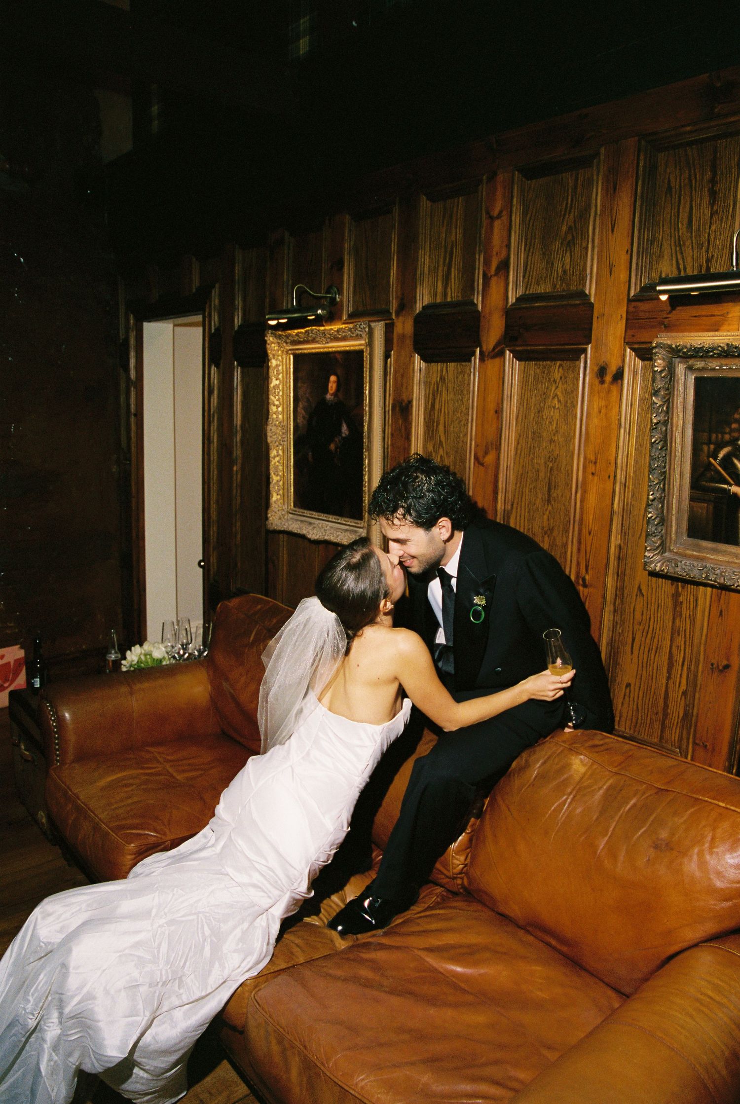 Bride and groom sharing a romantic kiss on a leather couch surrounded by framed portraits.