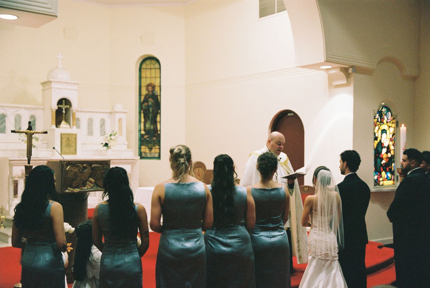 Bridesmaids in teal dresses stand at church altar during traditional Catholic wedding ceremony.