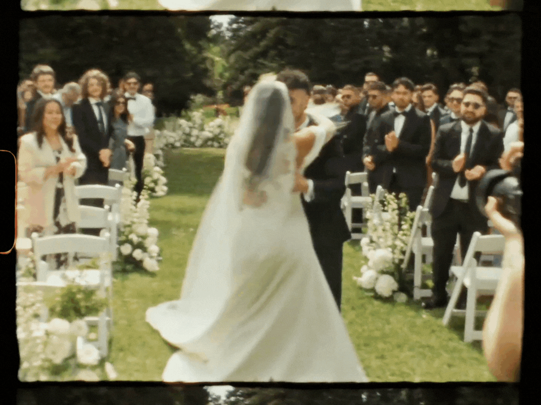 Bride in white gown and veil walks down outdoor aisle past guests at elegant garden wedding ceremony.