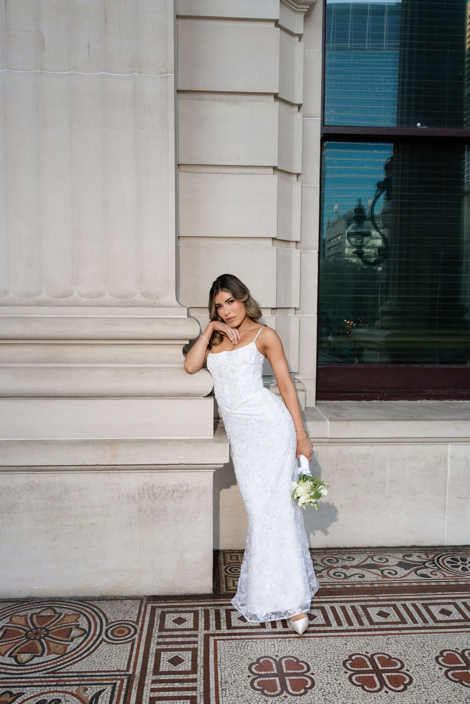 Elegant bride in white lace mermaid gown holding bouquet leaning against neoclassical stone building.