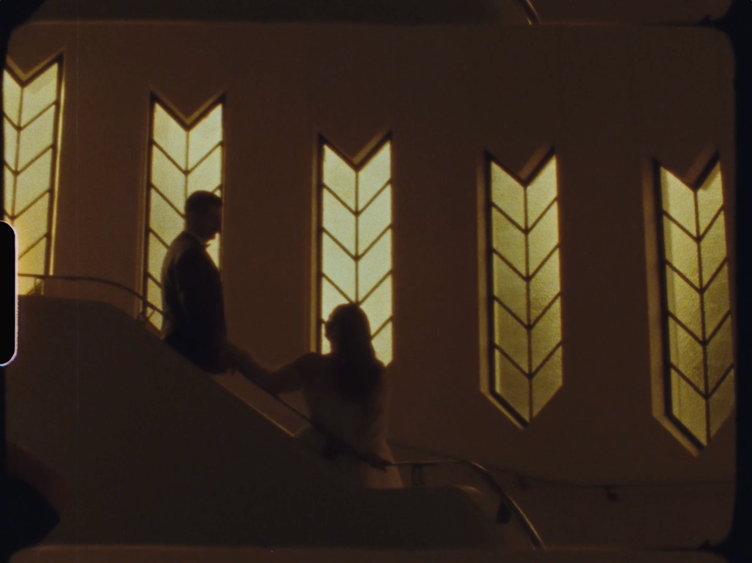 Silhouettes of a couple on stairs beside glowing arrow-shaped stained glass windows in a church.