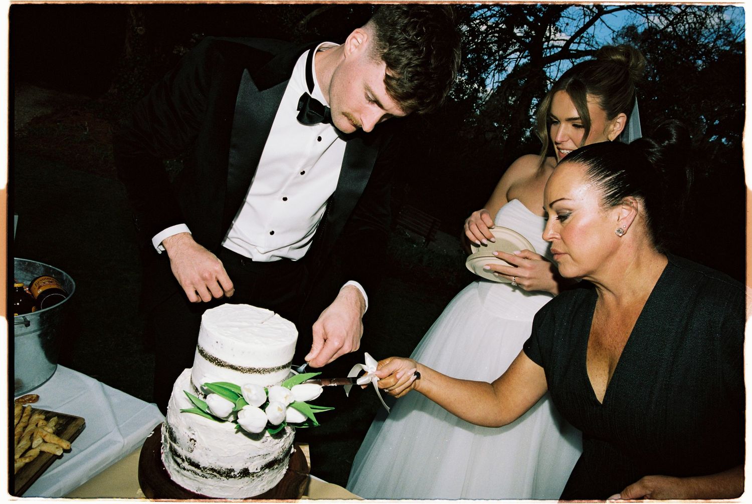 Bride and groom cut tiered white wedding cake decorated with greenery while guest watches closely.