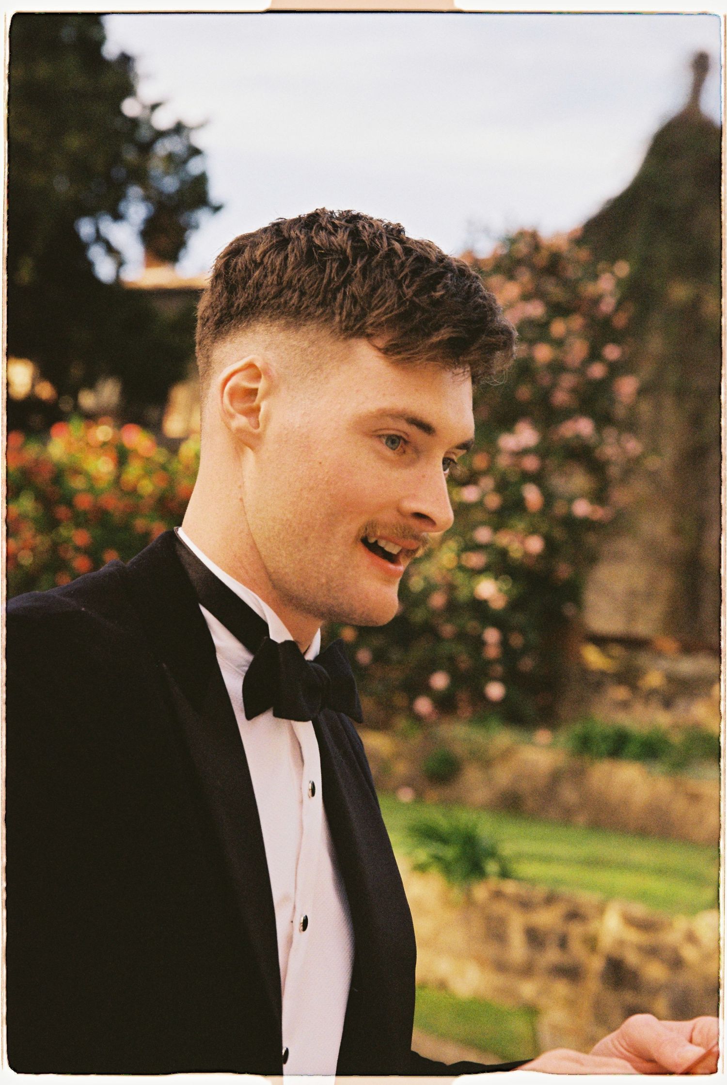 Young man in a black tuxedo with bow tie smiles in a garden with colorful roses in the background.