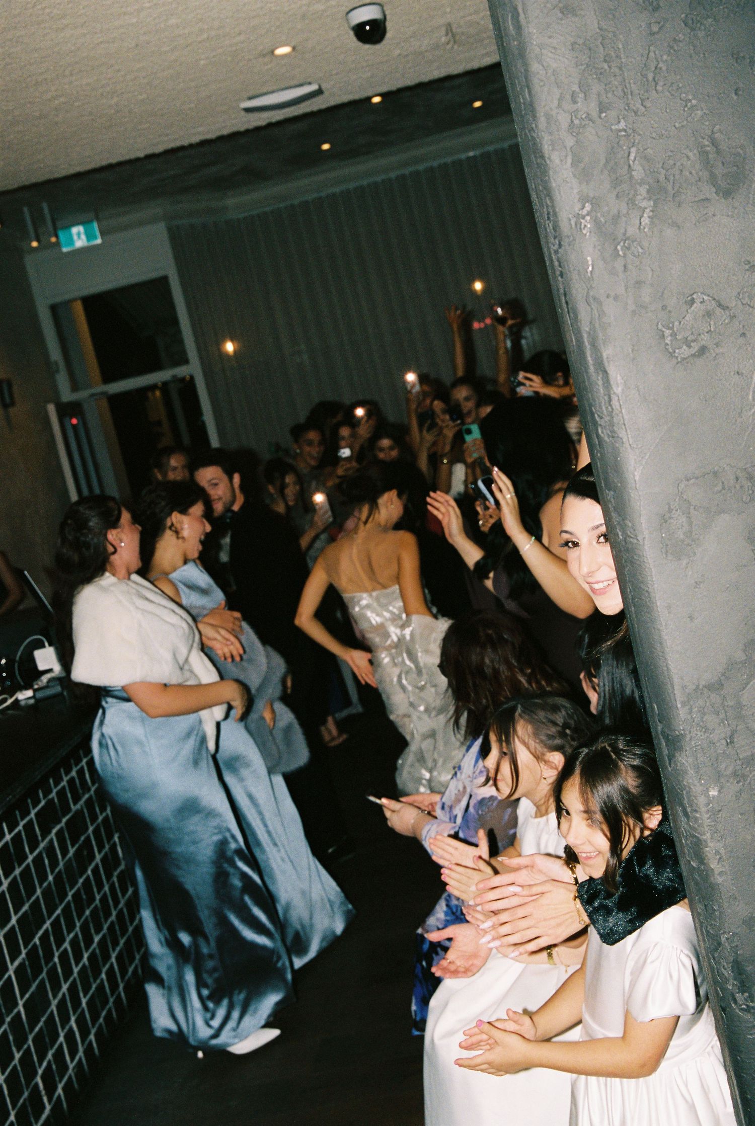 Guests in formal attire line a corridor at a wedding reception, holding phones up to capture the moment.