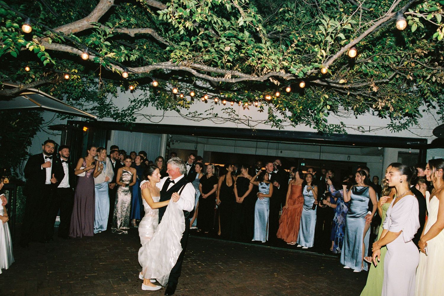Bride and groom share first dance outdoors under string lights and lush trees surrounded by wedding guests.