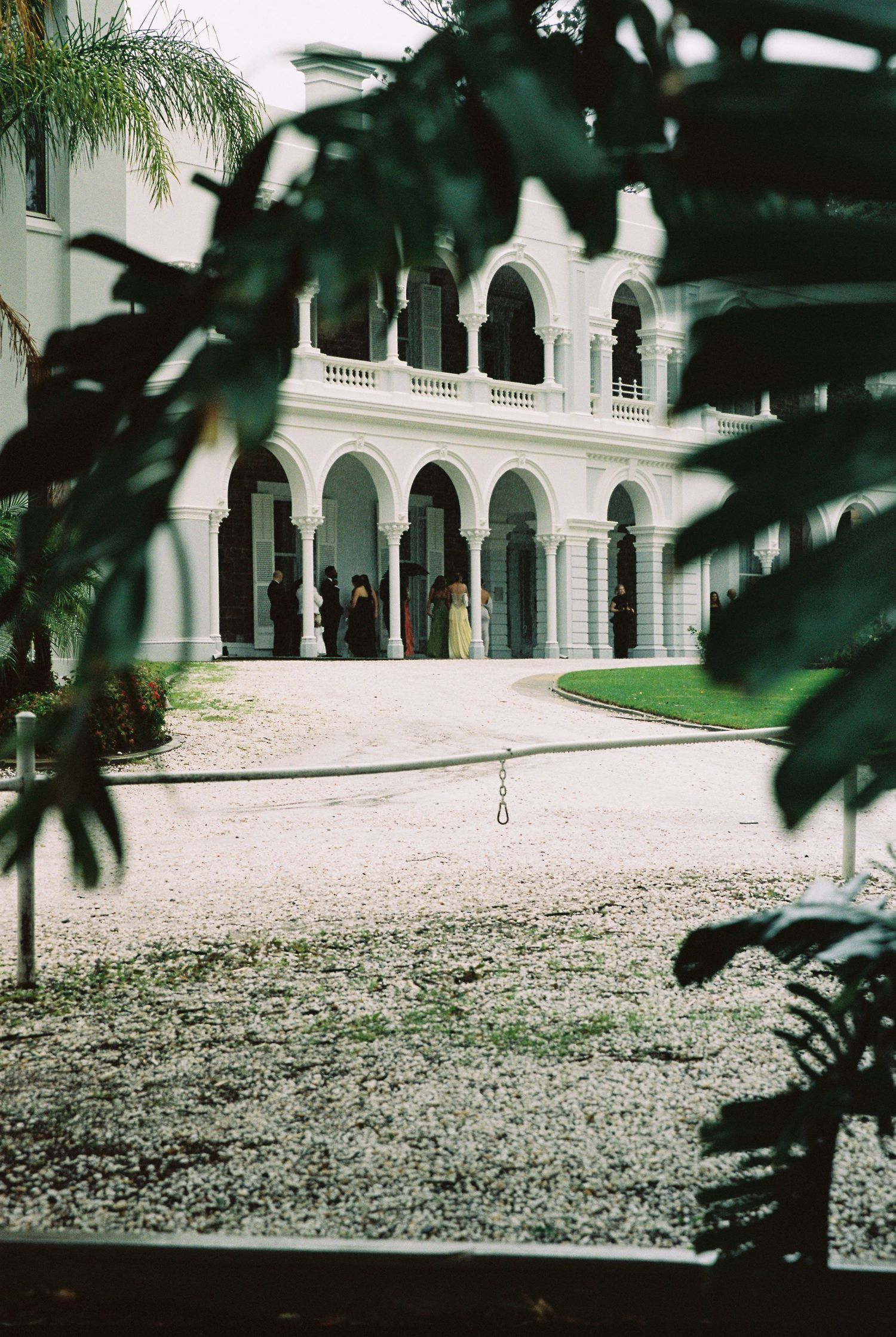 Elegant white colonial wedding venue with arched colonnades viewed through tropical foliage.