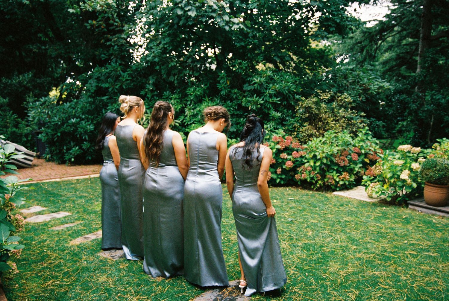 Four bridesmaids in matching silver satin gowns standing together in a lush garden setting.