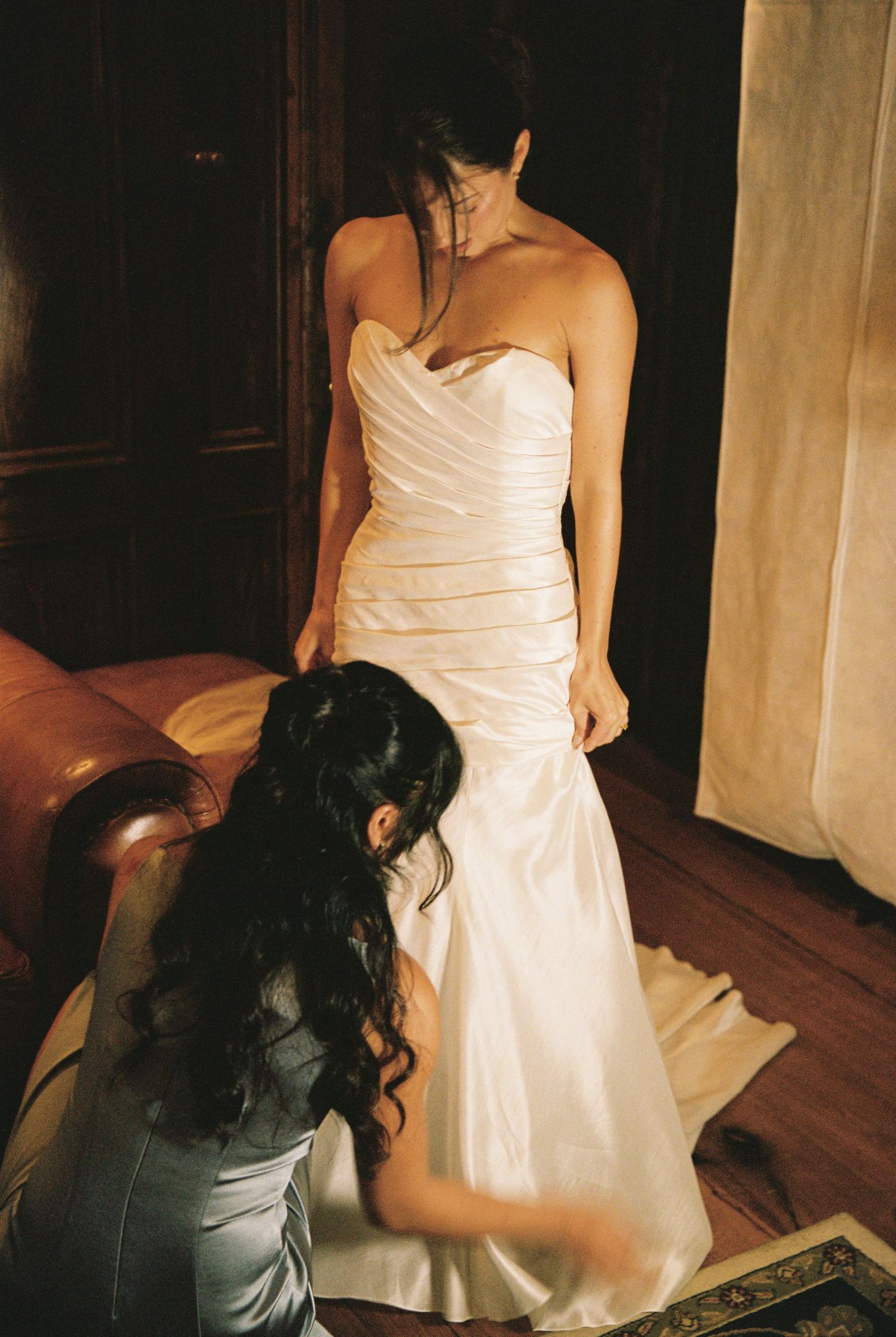 Bridesmaid helping bride into elegant strapless ruched white wedding gown in warmly lit room.