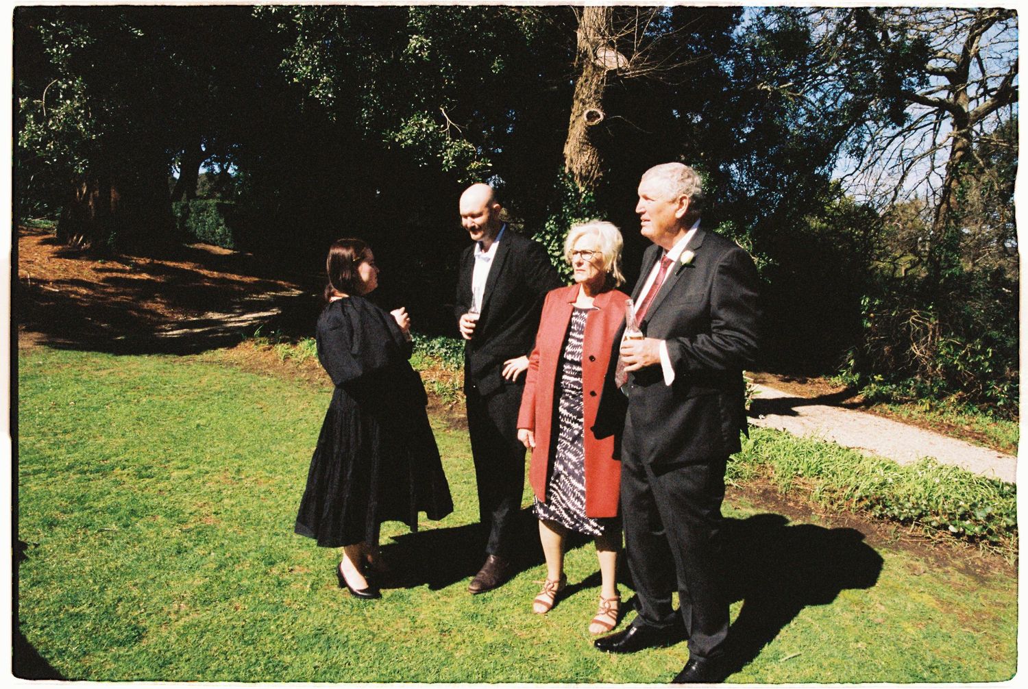 Four elegantly dressed guests conversing outdoors in a lush garden setting.