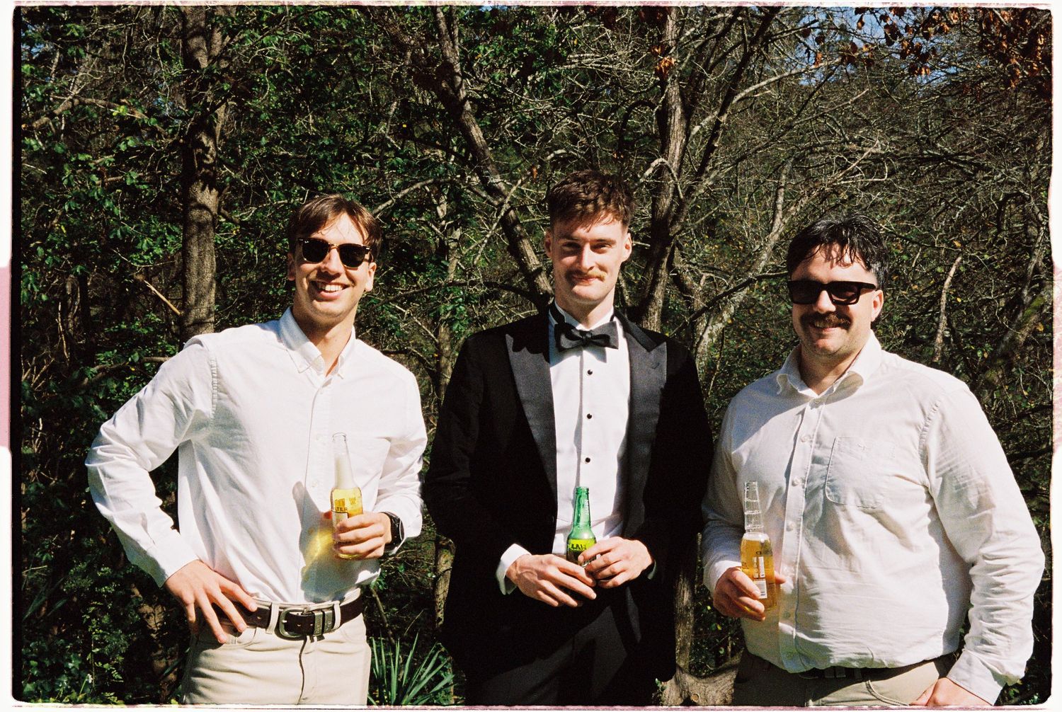 Three men in formal attire holding drinks outdoors, with two in white shirts and one in a black tuxedo, surrounded by trees.