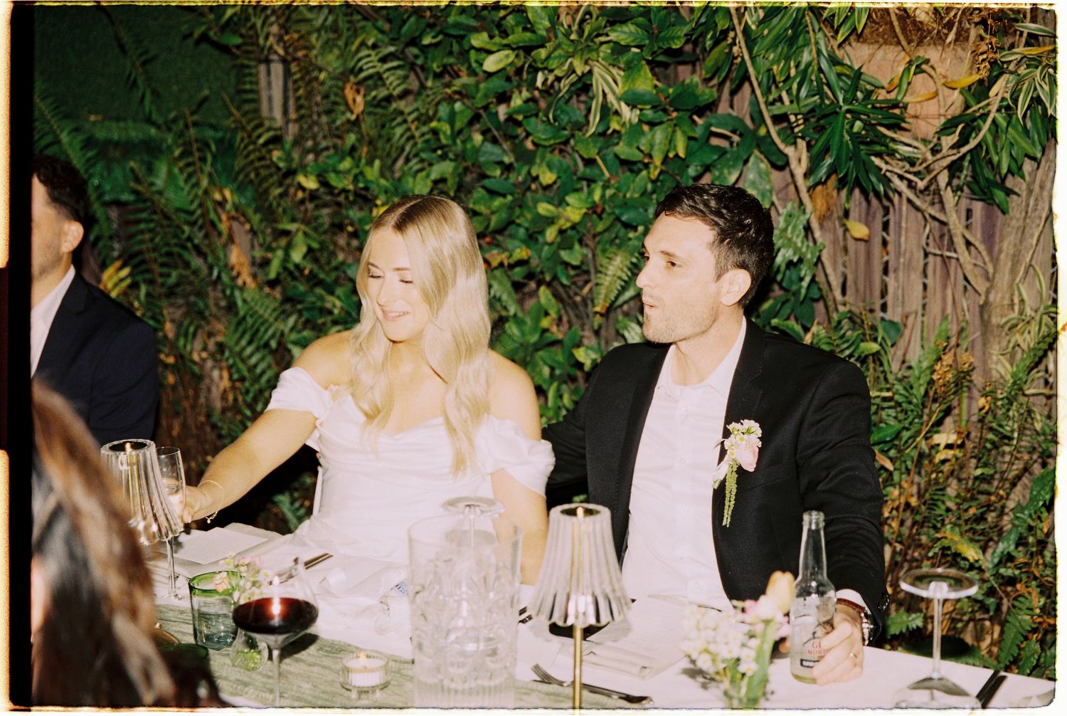 Bride and groom smiling together at wedding reception table surrounded by greenery.