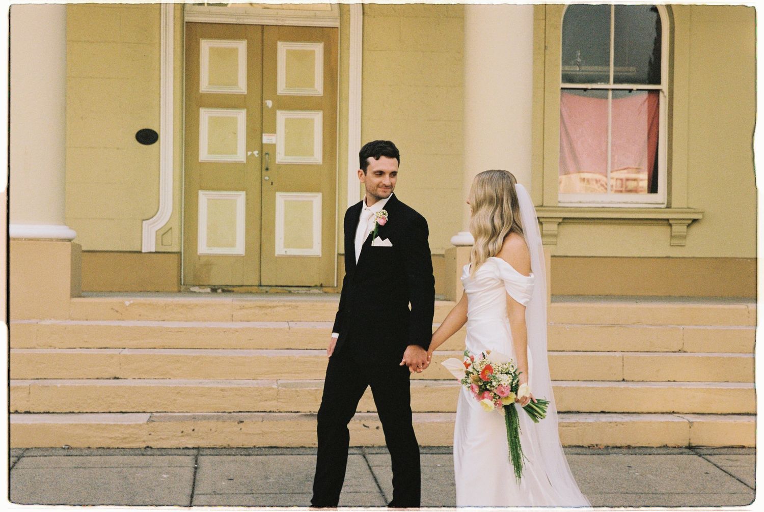 Bride and groom holding hands in front of a yellow building on their wedding day.