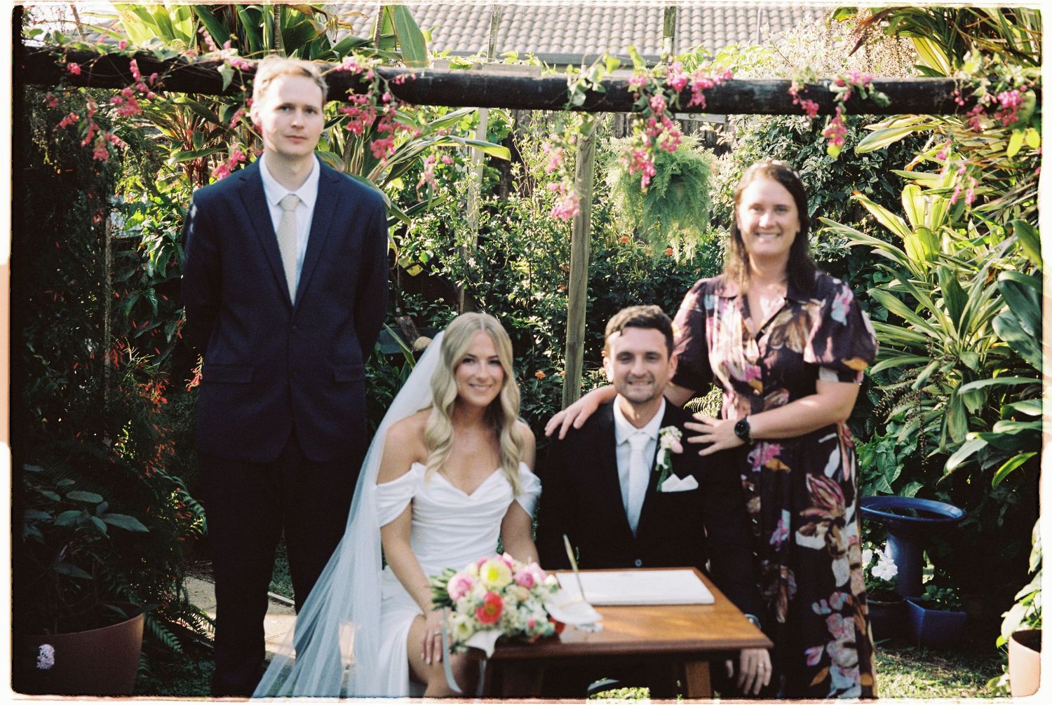 Newlywed couple signs marriage certificate with witnesses at outdoor garden wedding reception table.