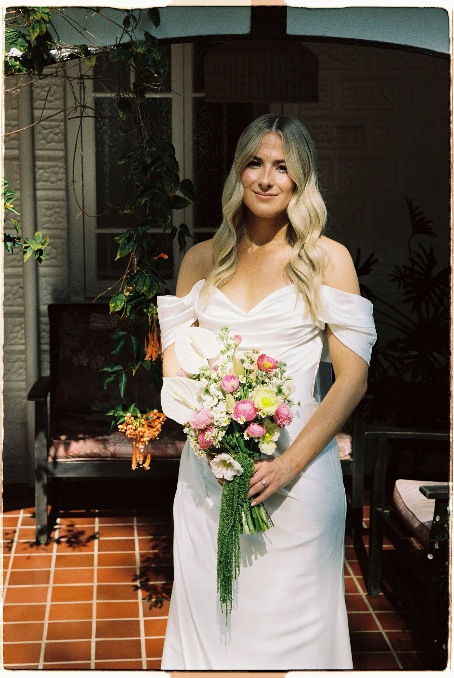 Smiling blonde bride in white off-shoulder wedding dress holding a colorful pink and yellow bouquet.