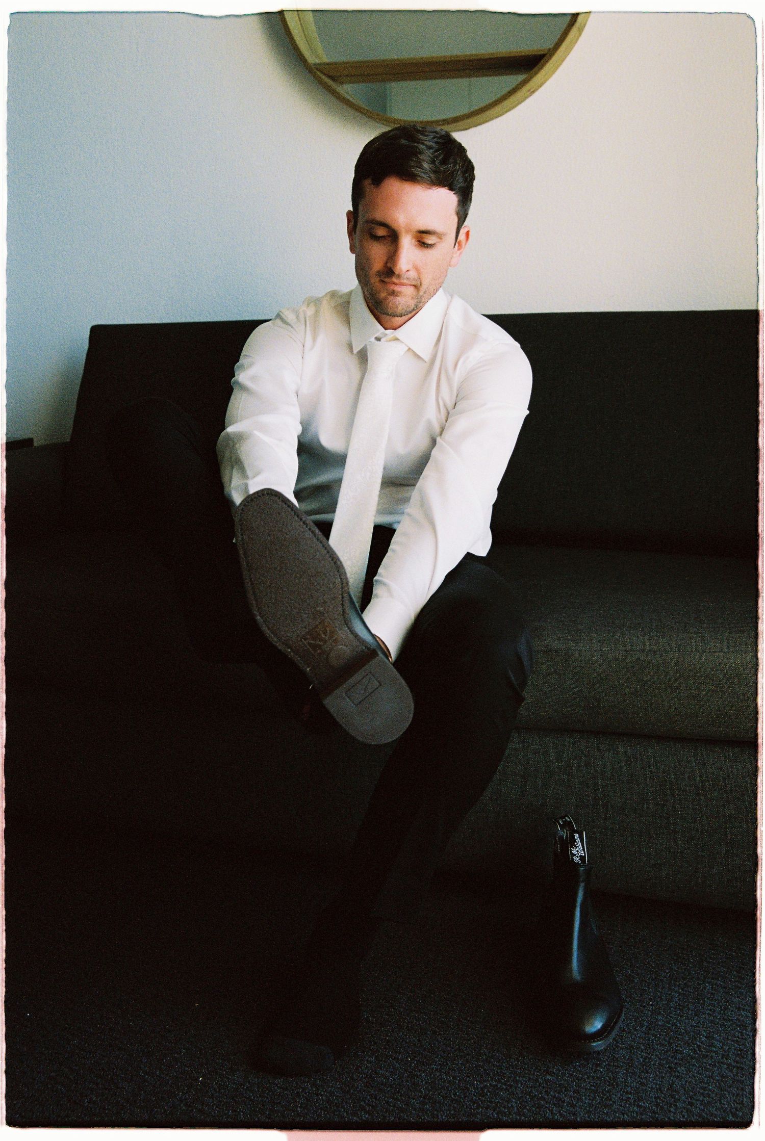Groom in white shirt and tie putting on dress shoes while seated on dark sofa.