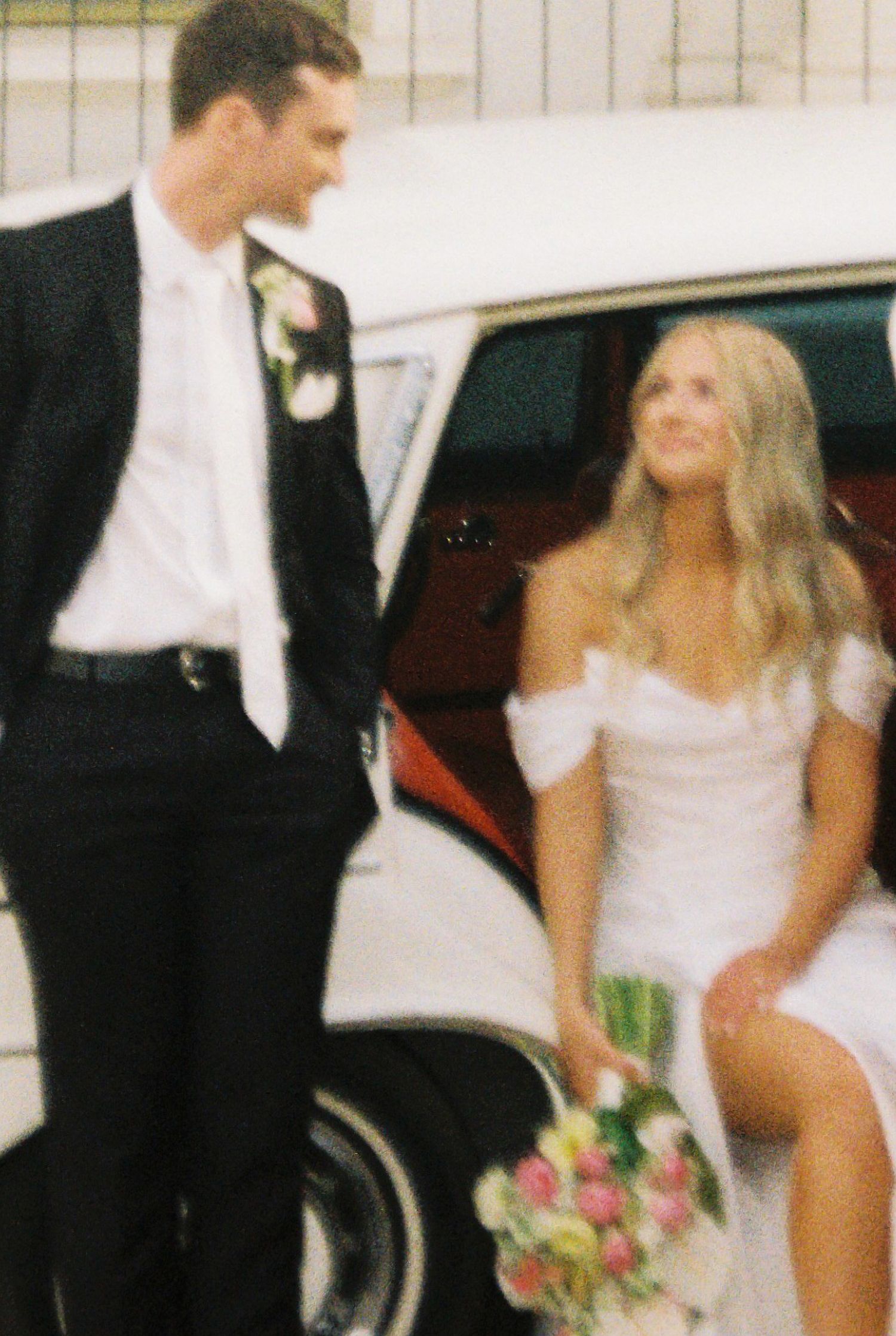 A bride in a white off-shoulder gown holds a floral bouquet beside a groom in a black suit near a vintage white car.