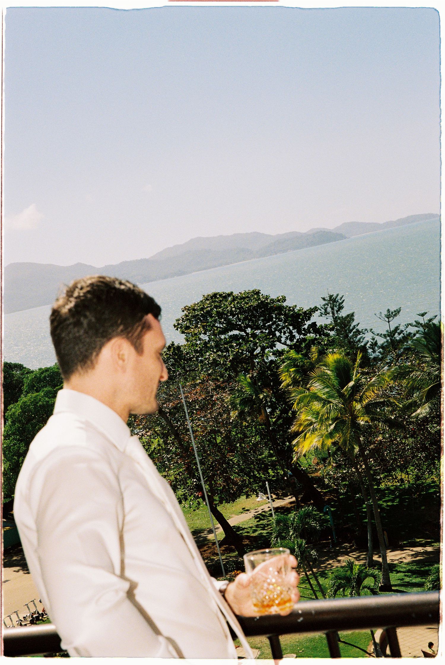 Man in white suit holding a whiskey glass on a balcony overlooking lush tropical greenery and ocean with mountains beyond.