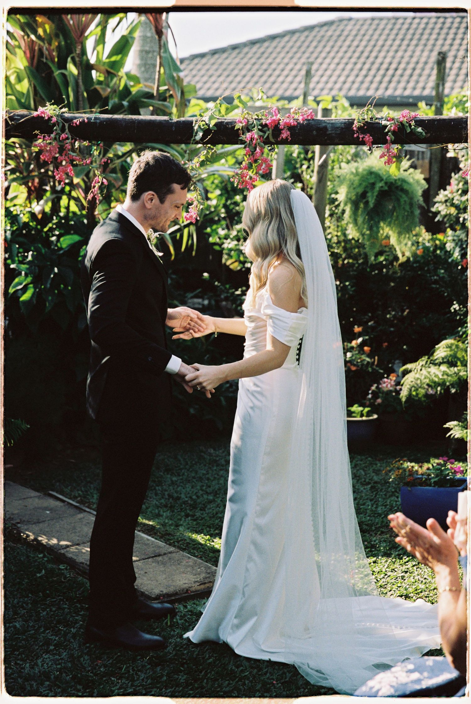 Bride and groom holding hands during outdoor garden wedding ceremony beneath floral arch.