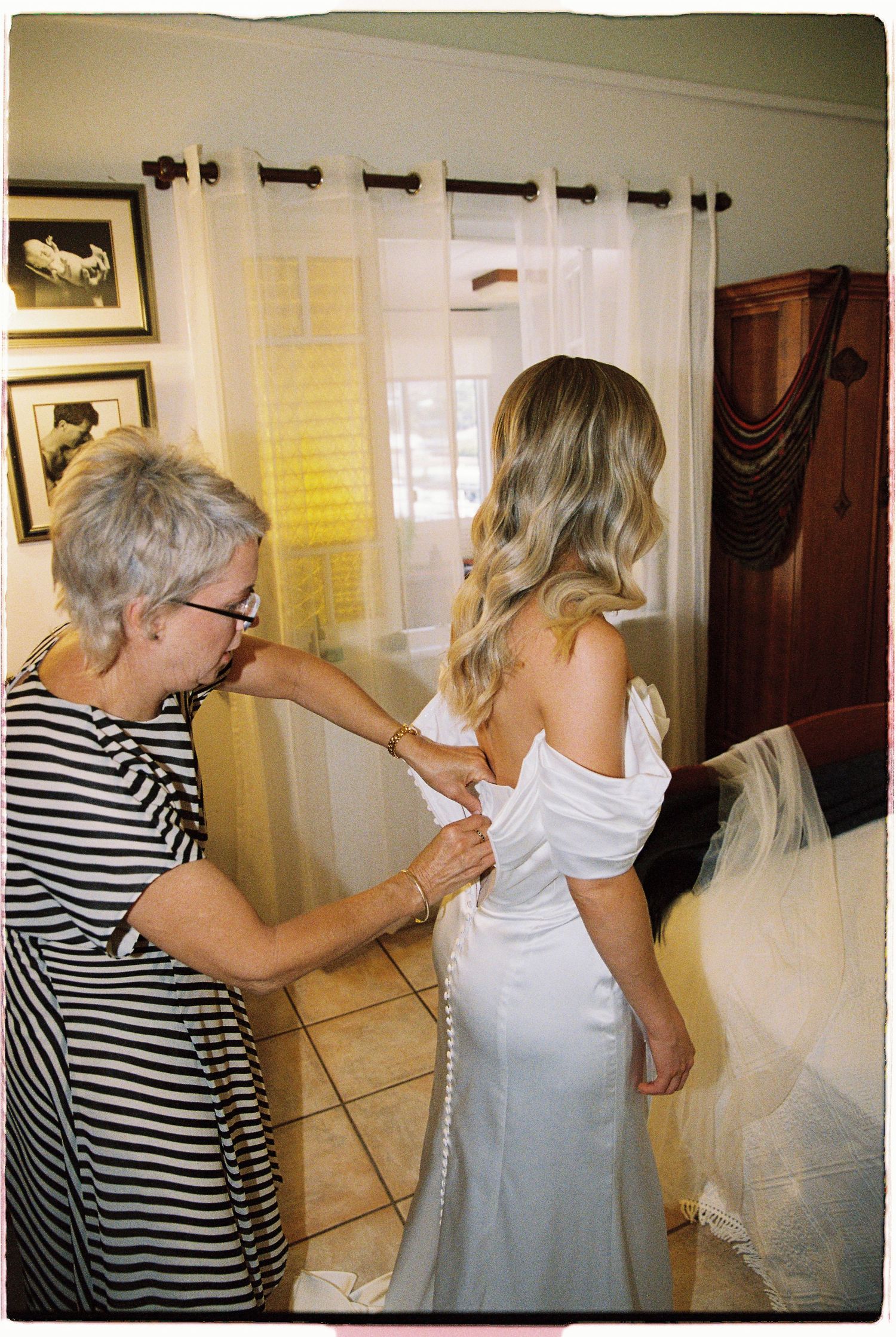 Mother helping bride button up white wedding gown in a warmly lit bedroom before the ceremony.