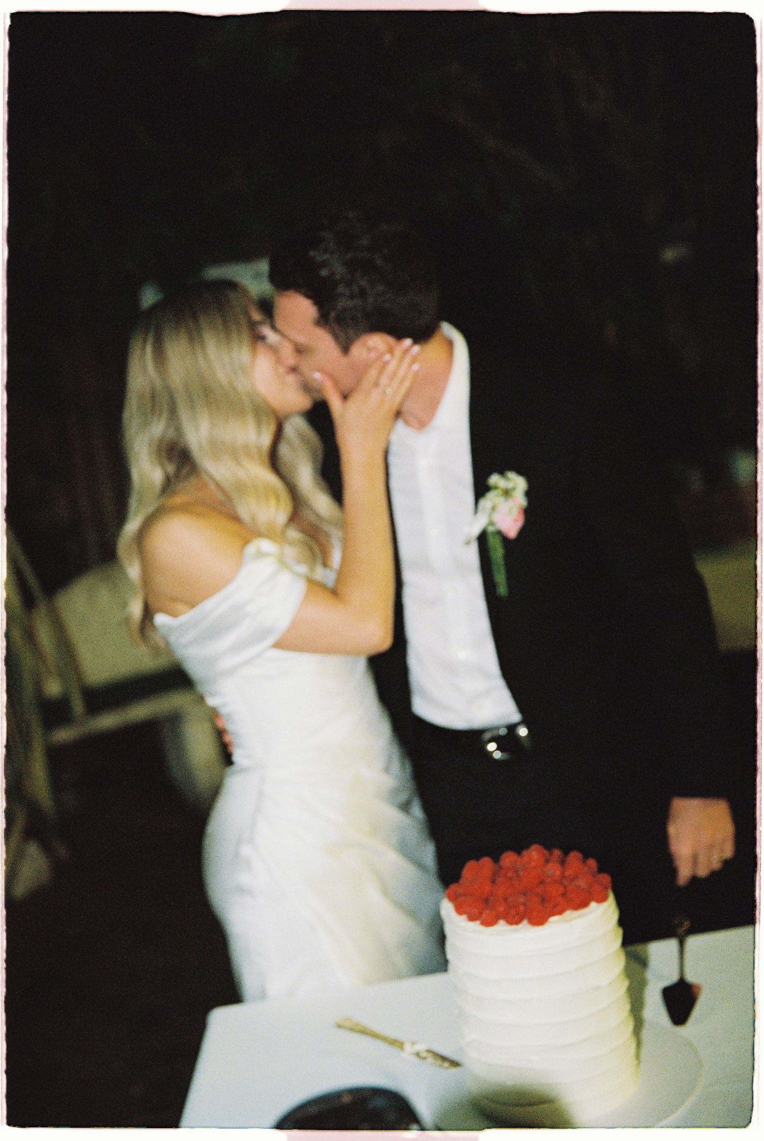 Bride and groom share a kiss while cutting their white wedding cake topped with fresh strawberries.