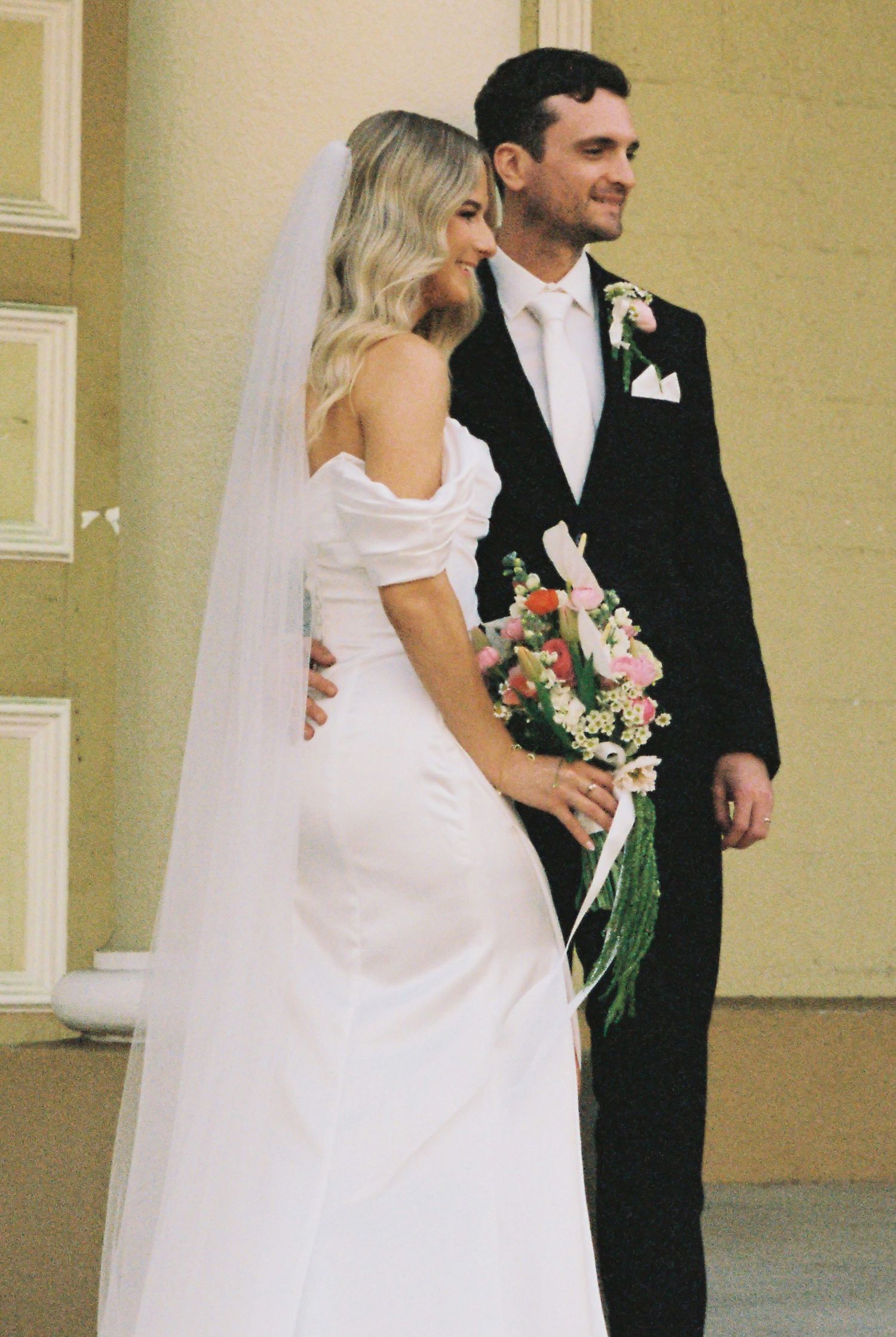 Bride and groom pose together outdoors, she holds a pink floral bouquet.
