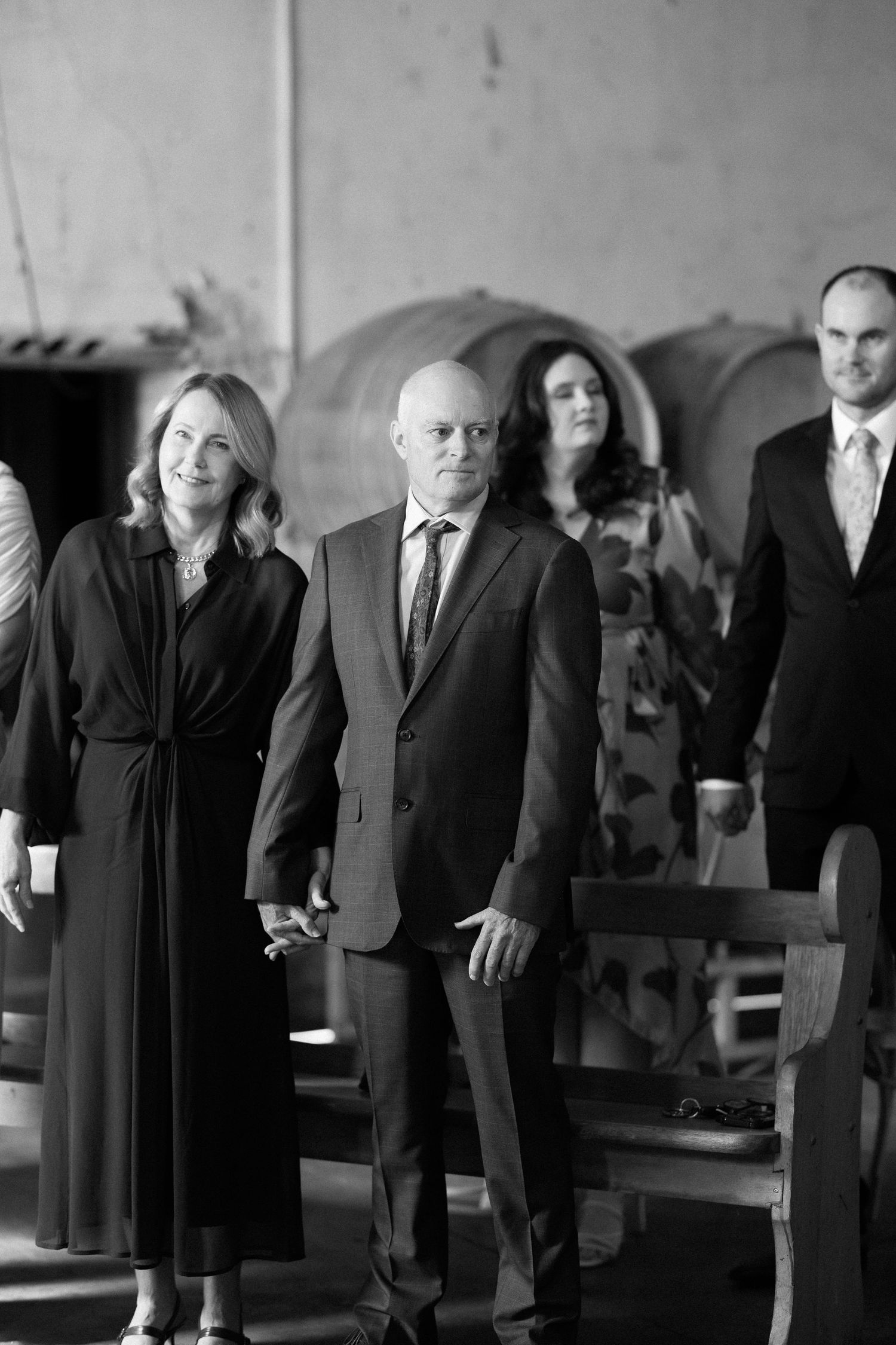 Elegantly dressed wedding guests holding hands walking through winery venue in black and white photo.