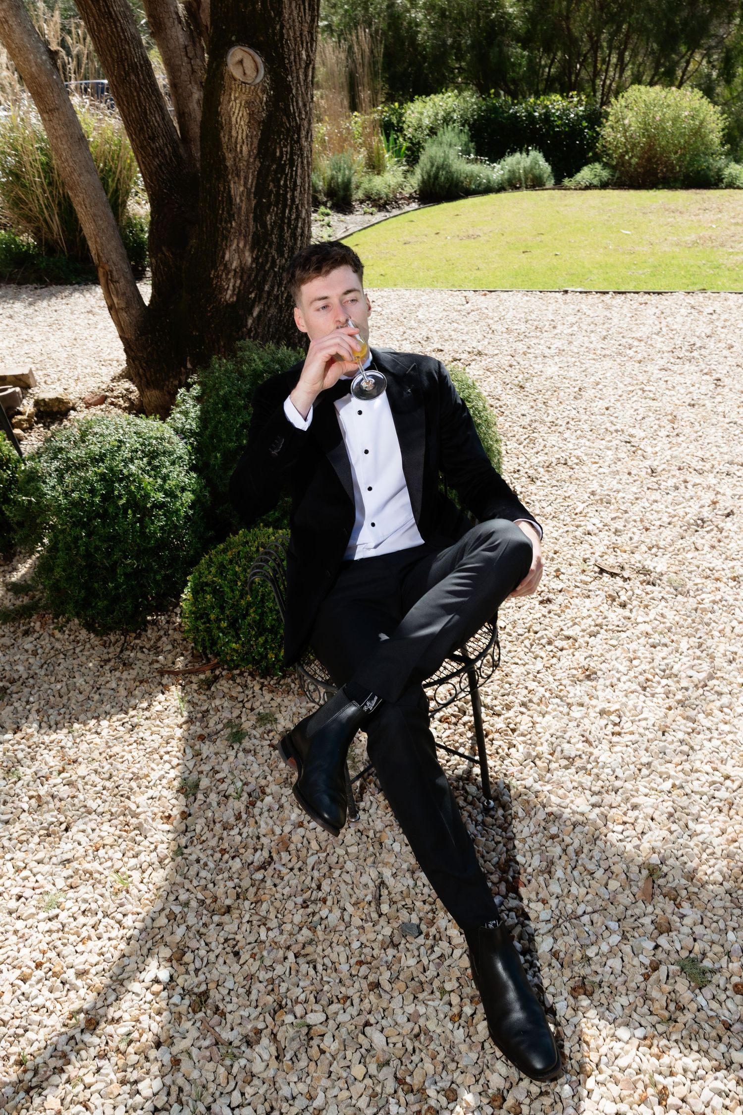 Groom in black tuxedo sitting on chair smoking near tree on gravel path.