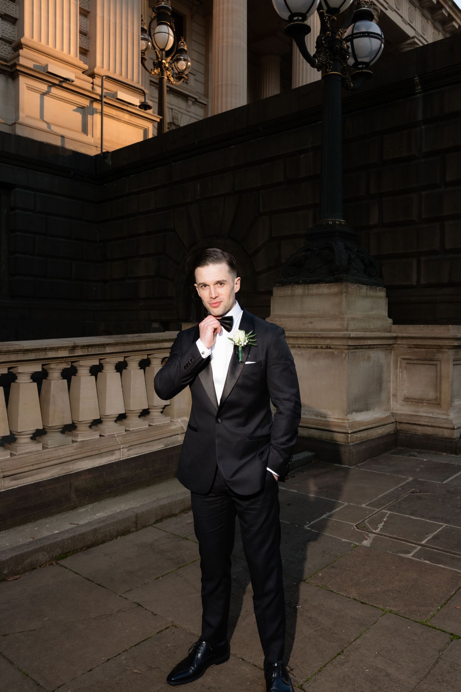 Dapper groom in black tuxedo with white boutonniere poses confidently outside elegant stone building.