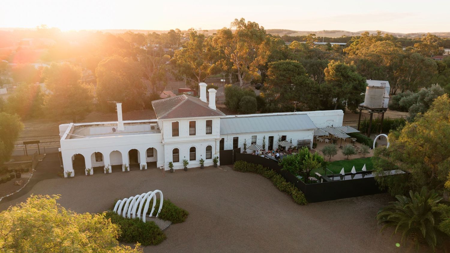 Aerial view of a white colonial-style estate surrounded by lush trees, bathed in golden sunset light.