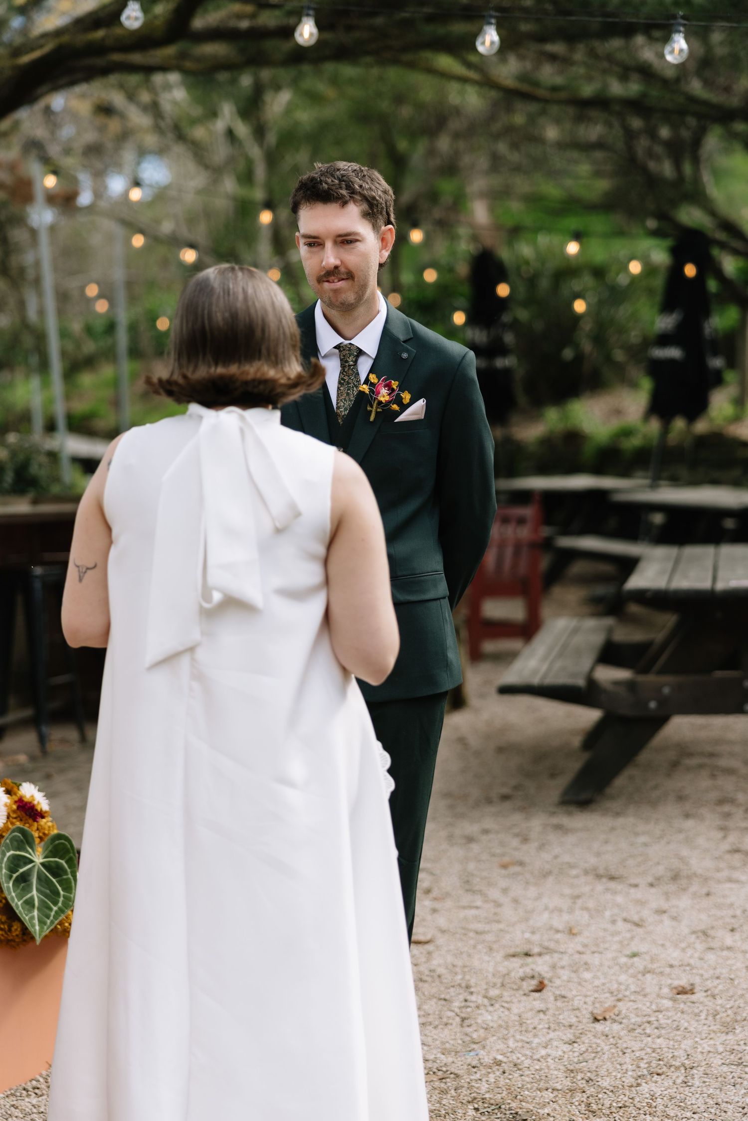 Groom in dark green suit smiles at bride in white dress during outdoor wedding ceremony with string lights.