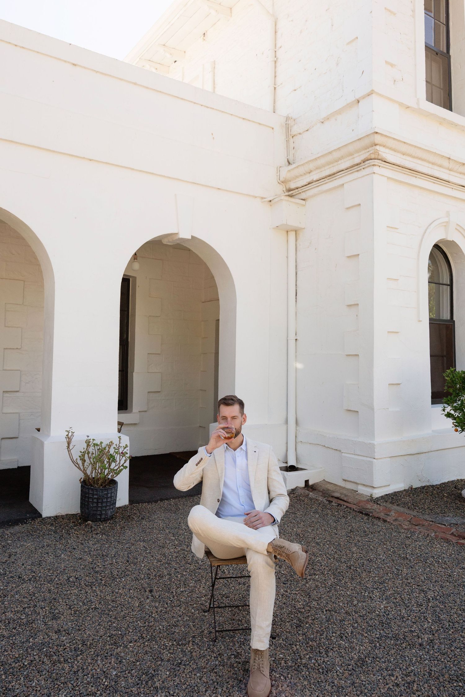 Dapper groom in beige suit sits confidently outside white arched heritage building.