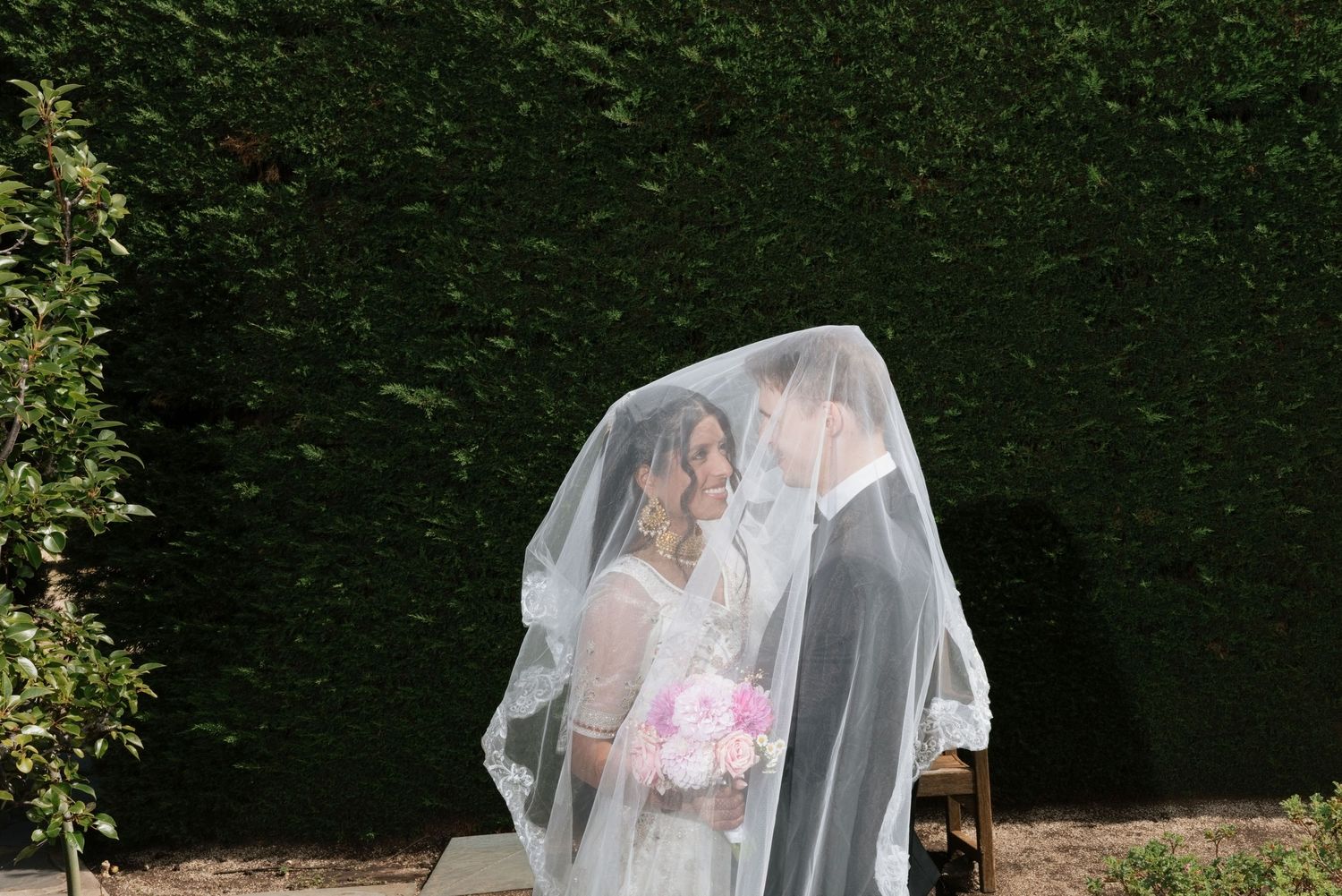 Couple sharing intimate kiss hidden under bridal veil in lush garden, bride holding pink floral bouquet.