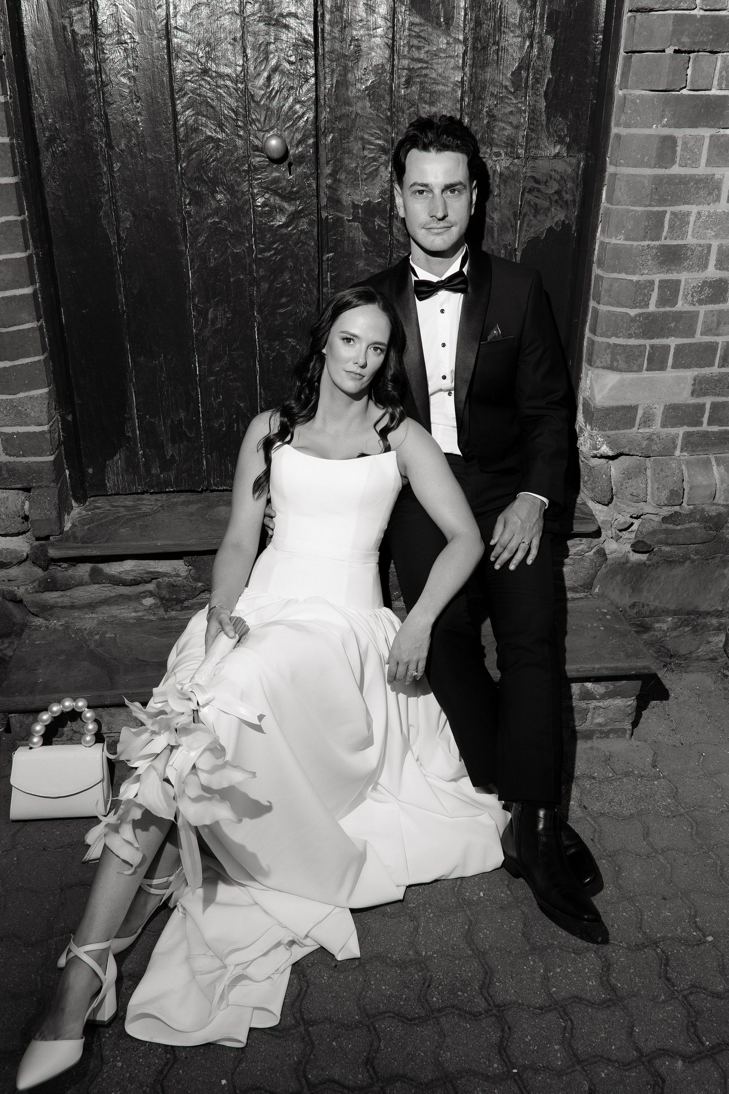 Bride and groom sitting together against rustic wooden door in black and white portrait.
