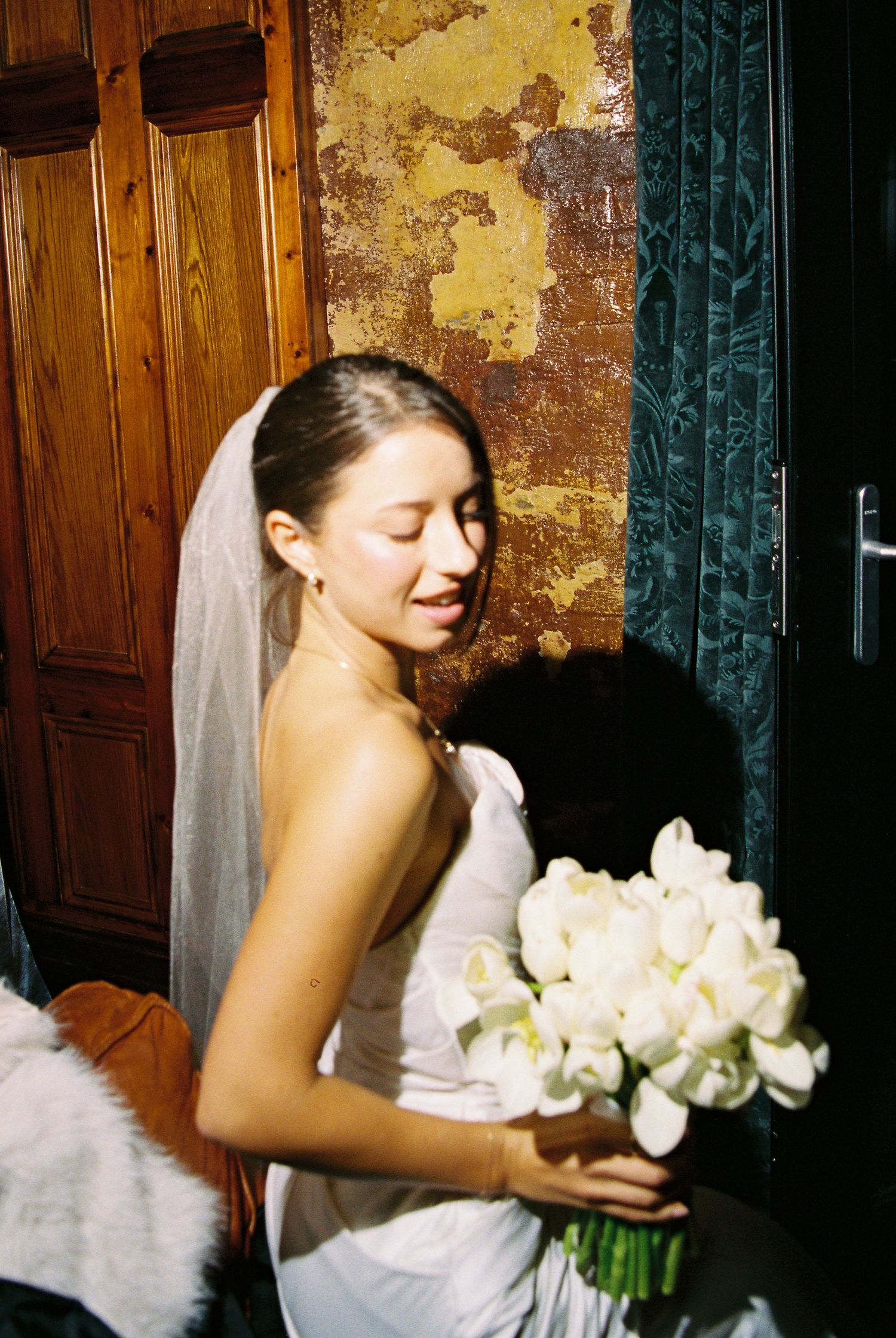Bride in strapless white gown holding white tulip bouquet with veil beside rustic peeling wall.