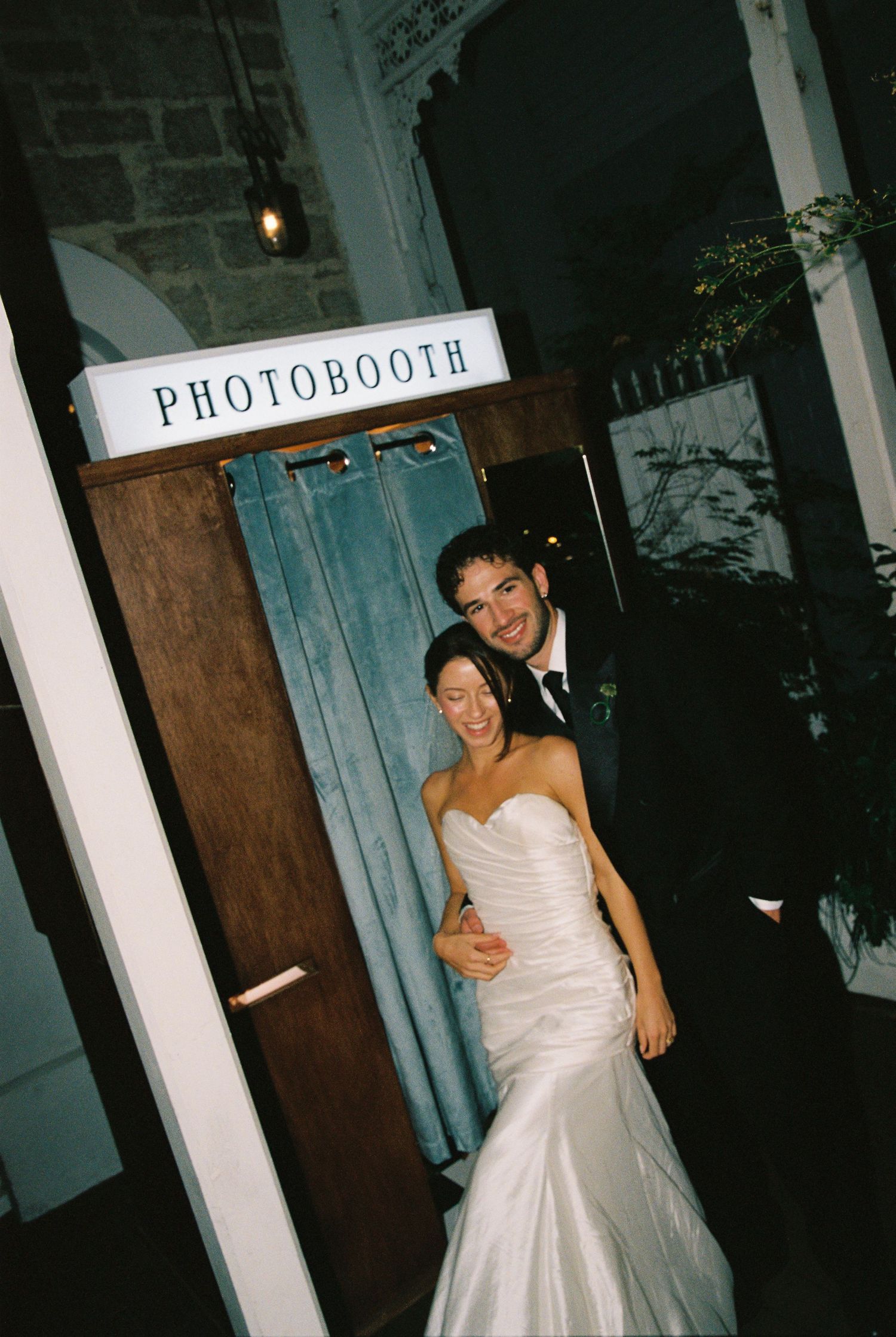 Bride and groom smile together outside a photobooth at their elegant evening wedding reception.